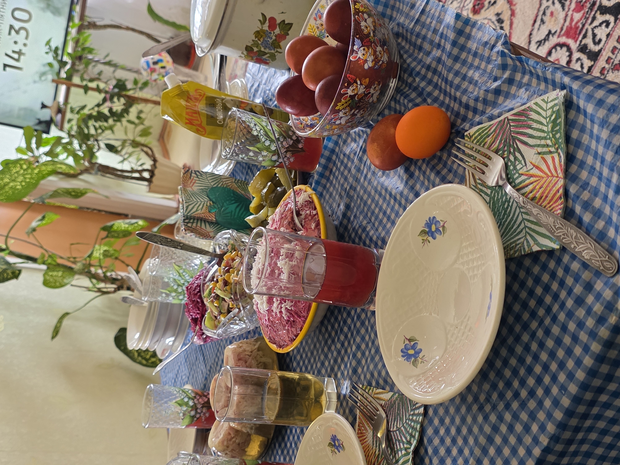 The image shows a table set for a meal, covered with a blue and white checkered tablecloth. A bowl of painted eggs sits on the table, along with a plate, a fork resting on a tropical-patterned napkin, and various dishes containing salad and other foods. A glass filled with red liquid is also visible. A potted plant is placed to the side of the table. The scene appears to be indoors. A television screen showing the time "14:30" is visible in the background. The photograph was taken in Chaikovsky, Russia.