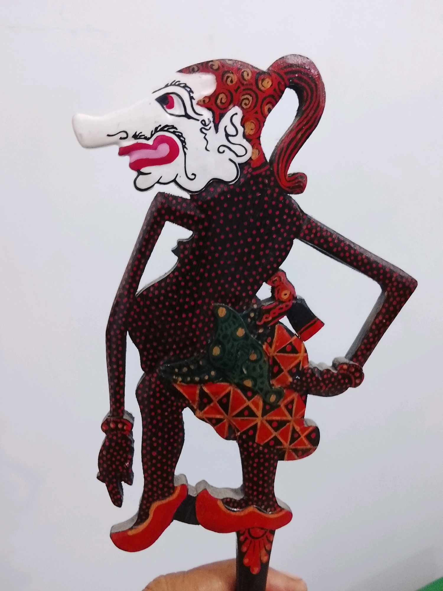 A wooden Wayang Golek puppet is held up against a white background. The puppet is elaborately decorated with red and black polka dots, red and gold swirls on its head and hair, and a patterned skirt. The puppet has a long, white nose, red lips, and painted red eyes with black eyeliner. Its hands are depicted in a stylized manner, and it wears red shoes. A person's hand is visible at the bottom of the image, holding the puppet's stick. The scene is likely taking place indoors, given the plain white background, though a precise location within Slawi, Indonesia, cannot be determined from the image alone.