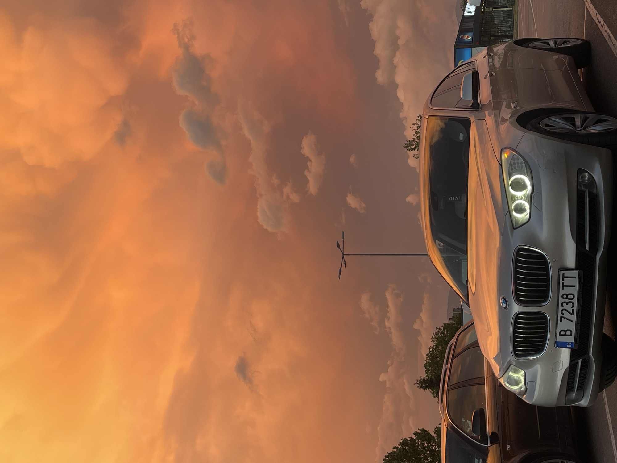 The image shows a silver BMW with the license plate "B 7238 TT," seemingly parked in Kazanlak, Bulgaria. The background is dominated by a dramatic sunset with orange and brown clouds. In the middle ground, there's a streetlight. Some trees and the corner of a building are visible in the upper portion of the frame.