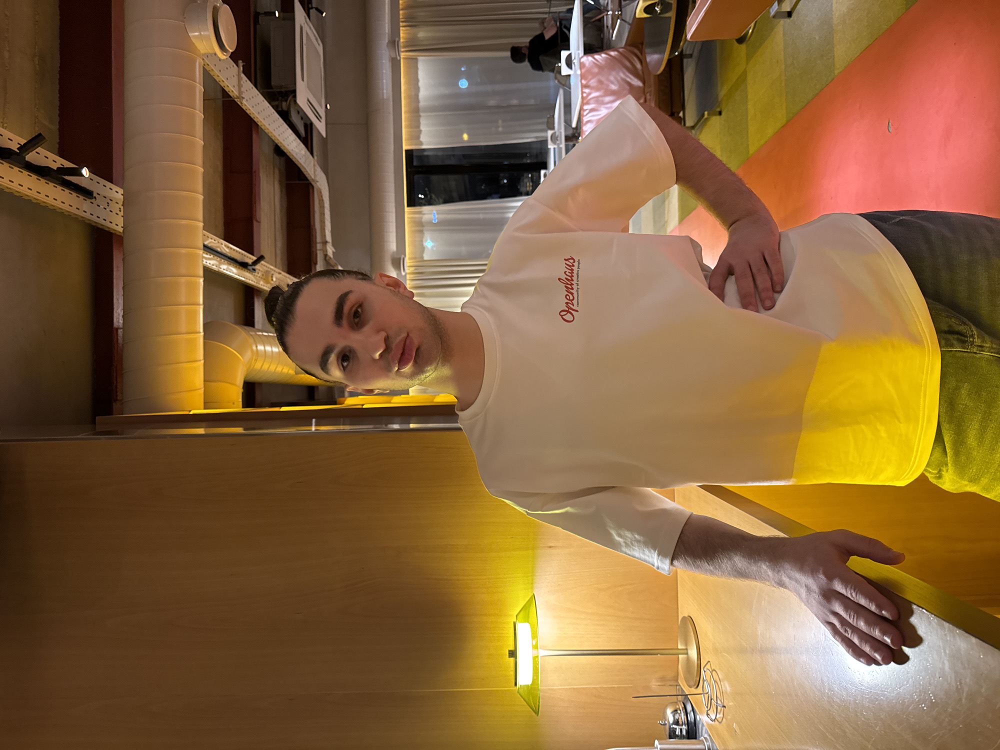The image shows a man with dark hair pulled back in a bun wearing a white t-shirt with the word "Orenhaus" written in red on the chest. He is standing in what appears to be a restaurant or bar setting. His left hand is resting on his lower back and the right hand is on a reflective surface.
Behind the man, there is a restaurant area with tables and chairs, with a person visible in the background. The lighting is a mix of natural and artificial. There is a bright yellow lamp on the counter in front of the man.
Architectural details include exposed ductwork and piping on the ceiling and walls, suggesting an industrial or modern design. The floor has alternating stripes of yellow and red.
The specific location in Yerevan, Armenia, cannot be determined from the given image.