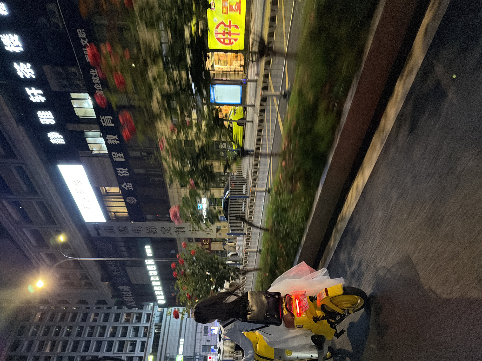 A nighttime shot captured in the Chongqing High-tech Zone, China, features a person riding a yellow scooter down a street lined with buildings. The street is adjacent to a sidewalk with vegetation and red flowers. Buildings on the left side of the road display vertical signs in Chinese. A streetlamp emits a bright glow. The scooter rider is facing the direction of travel along the street, slightly angled toward the right side of the frame.
