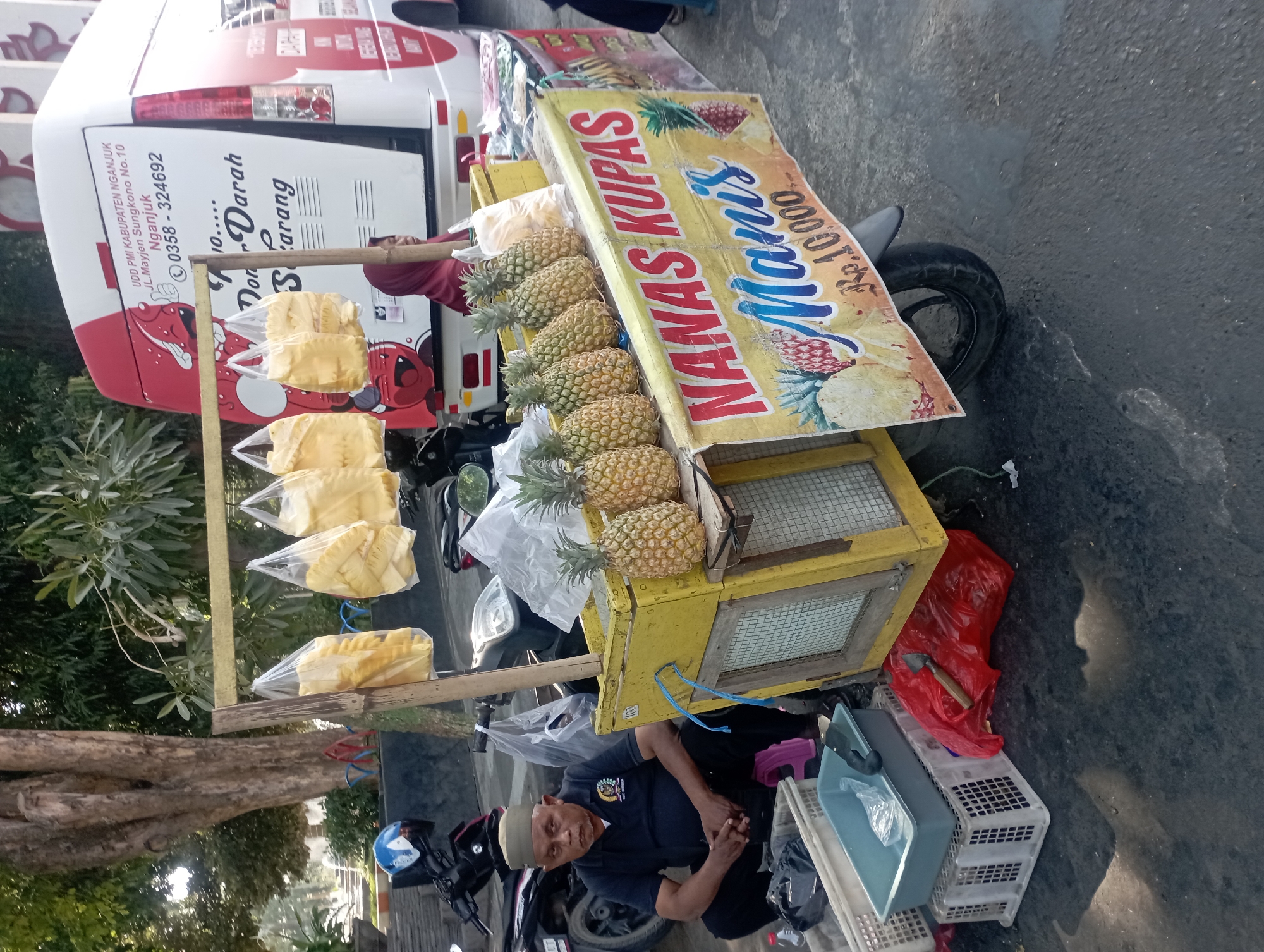 In Nganjuk, Indonesia, a vendor sits in front of a mobile cart selling pineapples. The cart is yellow and adorned with a banner that reads "NANAS KUPAS Manis Rp.10.000". Several whole pineapples are displayed on the cart, and bags of what appear to be pre-sliced pineapples hang from a wooden frame above. A man wearing a dark blue shirt and a light brown cap sits on a pink stool to the left of the cart, looking towards the viewer. Behind the cart, a white and red van with text in Indonesian, including "UDD PMI KABUPATEN NGANJUK" and a phone number, is partially visible. The ground is paved, and there are several plastic crates and a red plastic bag near the vendor. Motorcycles are parked in the background, suggesting a street-side location. The scene is outdoors, with trees and foliage visible in the background, indicating it is daytime.