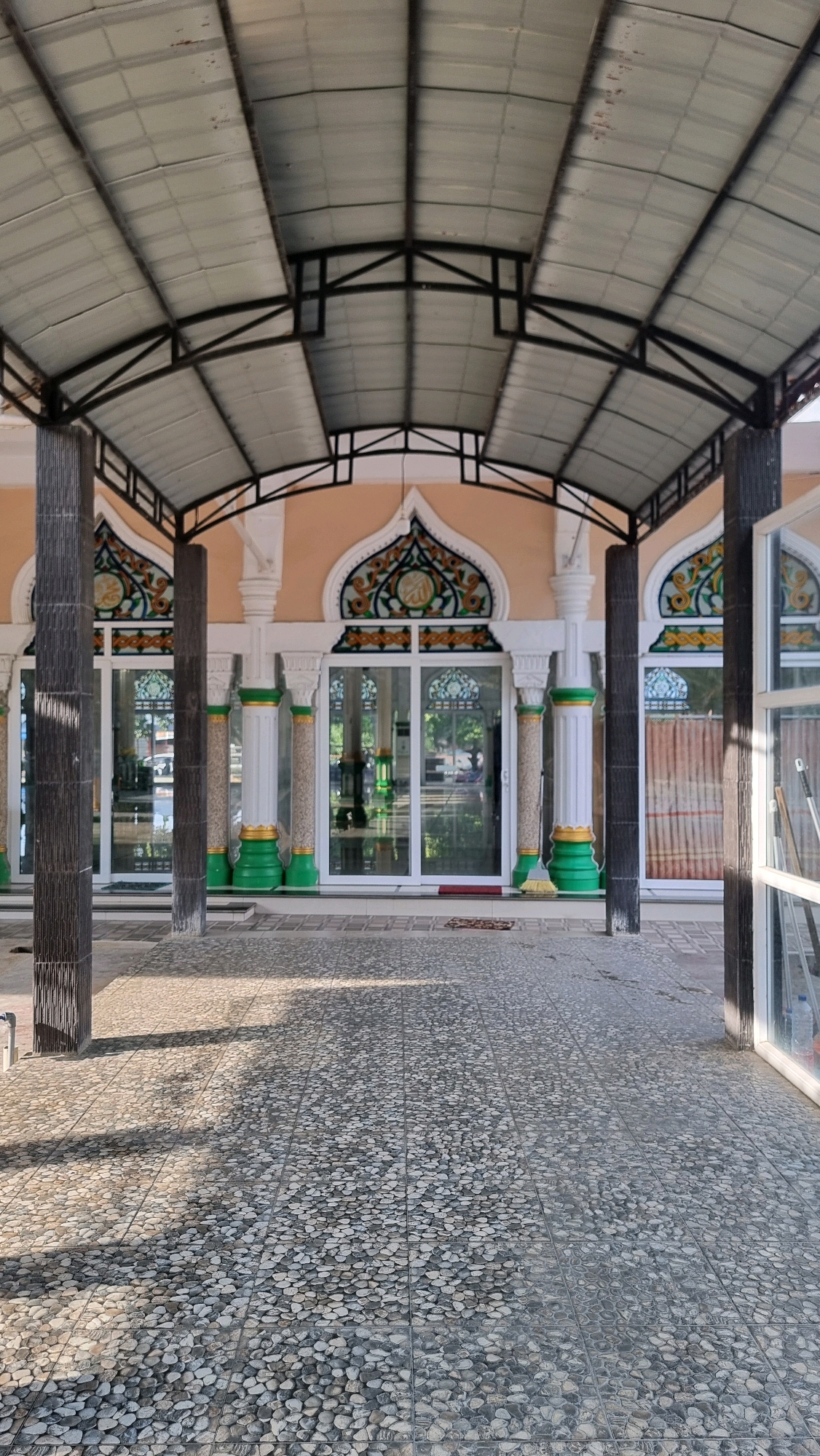 The image depicts the entrance to a mosque in Blangrambong, Indonesia. The building features a light orange facade with arched windows adorned with intricate green, gold, and blue decorative patterns. White columns with green and gold accents support a dark, corrugated metal roof supported by a metal framework. The ground in front of the mosque is paved with small, grey and white pebbles arranged in a tiled pattern, with shadows from the roof casting geometric shapes. Several tall, dark columns stand at the entrance, framing the view into the mosque's interior through large glass doors. Inside, the mosque appears to have a spacious interior with visible furniture and decorative elements. A broom leans against a column near the entrance, and a red doormat is visible. The overall impression is of a well-maintained religious building in an outdoor setting.