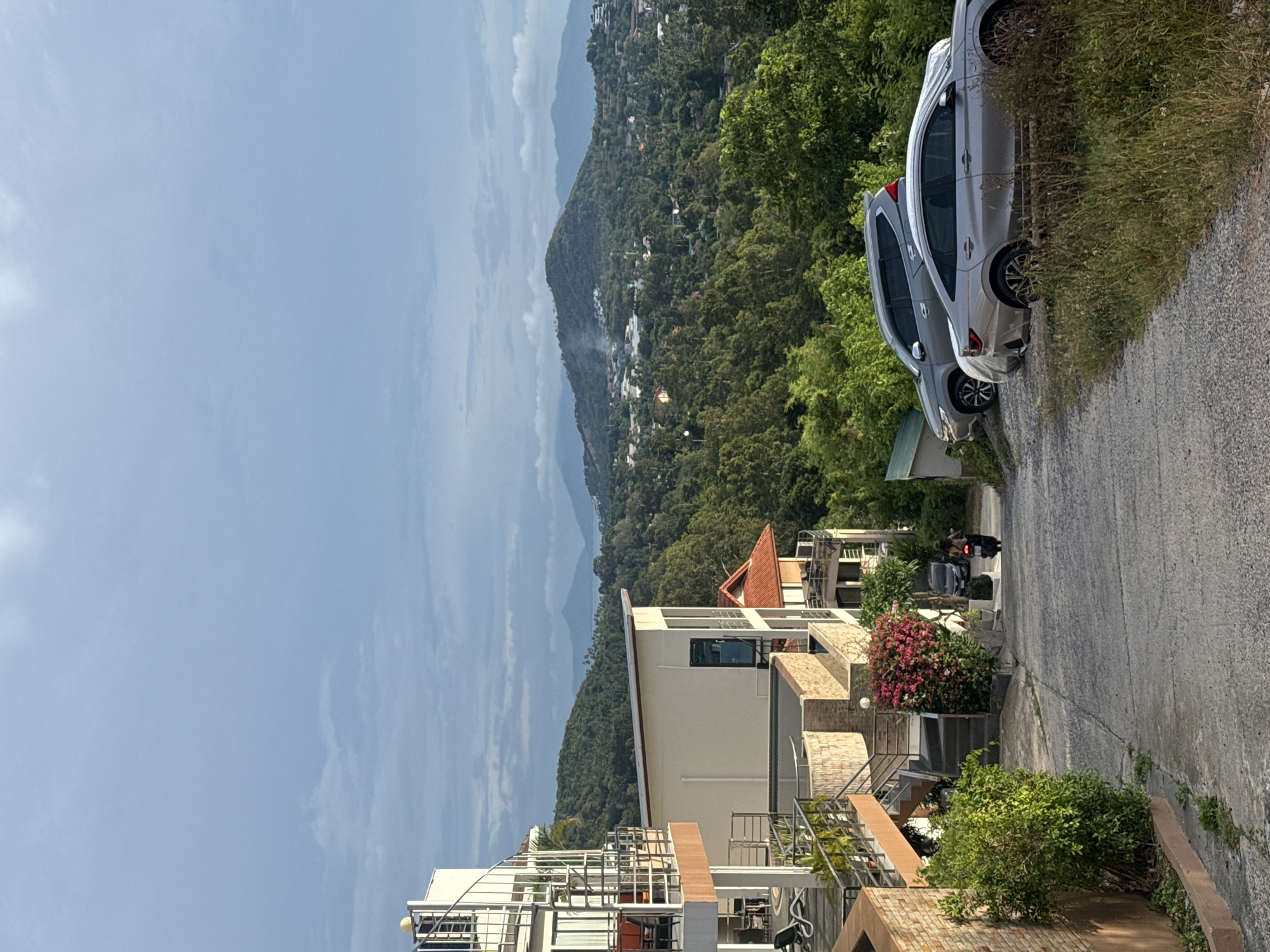 In Ko Samui District, Thailand, a paved road winds downhill, flanked by lush green foliage and modern multi-story buildings with balconies and spiral staircases. Two cars, a silver sedan and a grey SUV, are parked on the right side of the road. Further down, a person rides a motorcycle away from the camera, with another person walking beside them. In the background, rolling hills covered in trees extend towards a hazy horizon, with a village nestled amongst them and mountains visible in the distance. The sky is overcast with a pale blue hue.
