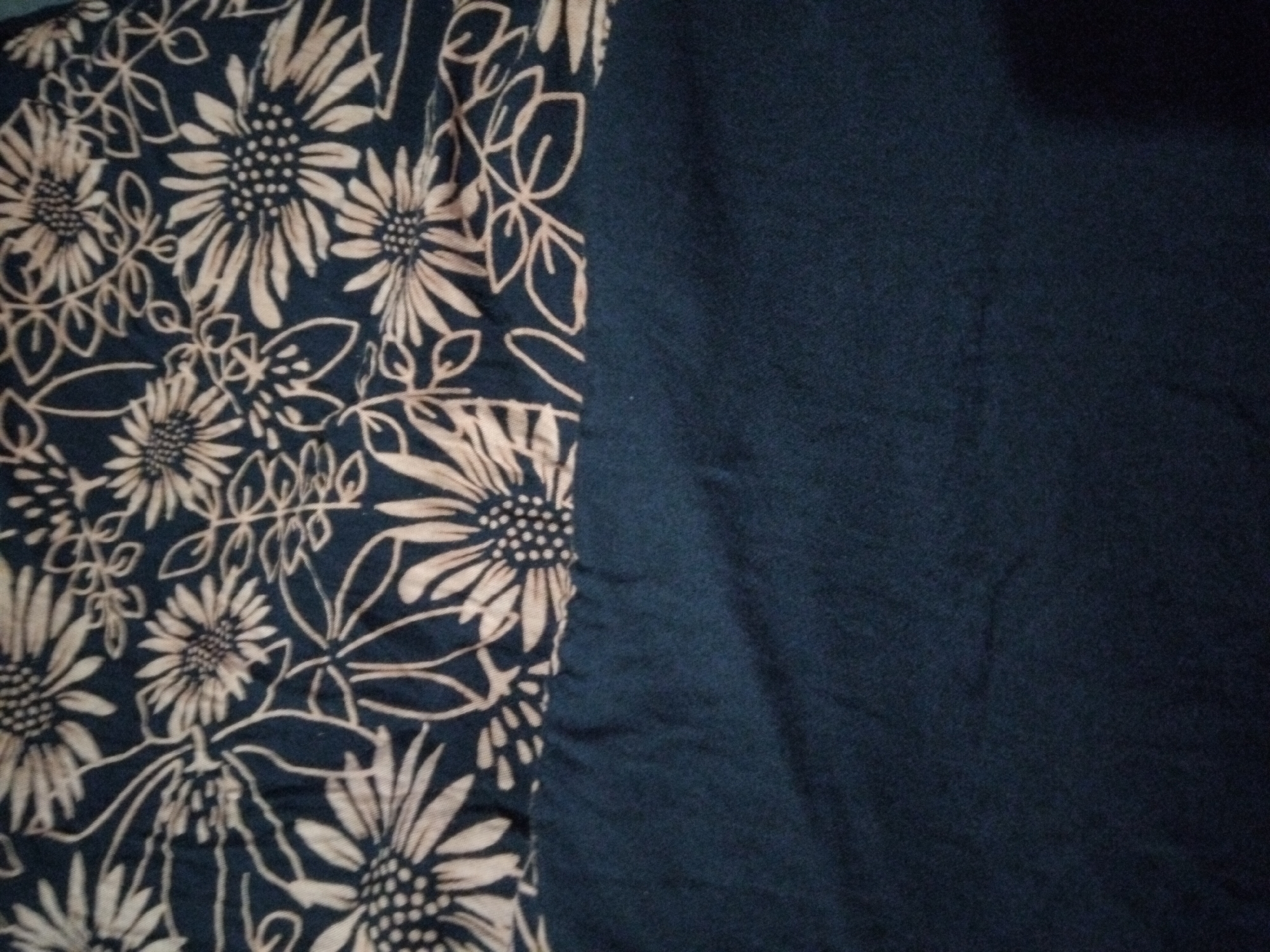 The image shows a close-up view of a dark blue fabric with a beige floral pattern. The pattern consists of stylized flowers with ray-like petals and some swirling vine-like elements. The fabric appears to be a garment or textile. The bottom portion of the image shows a solid, plain dark blue fabric. The scene is not indicative of any specific location in the city of Kepuh Kiriman, Indonesia, as it focuses solely on the texture and pattern of the fabric. There are no people, actions, or environmental elements visible in the image.