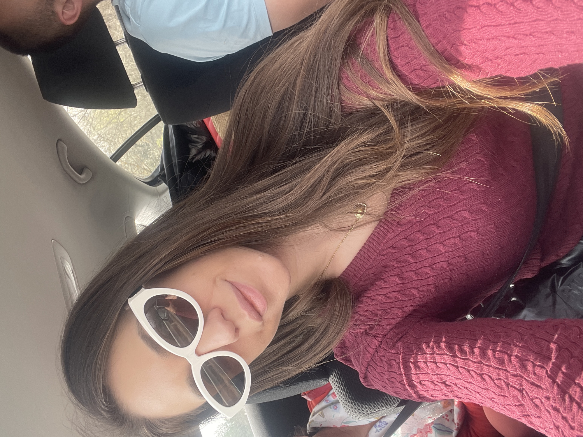 A selfie taken inside a car. A fair-skinned woman with light brown hair and white framed sunglasses is wearing a maroon cable knit sweater. The car appears to be parked, indicated by the headrest in view and the outdoor scenery visible through the rear window. The location, Kiosevtsi, Bulgaria, is not discernable from the visible scene.