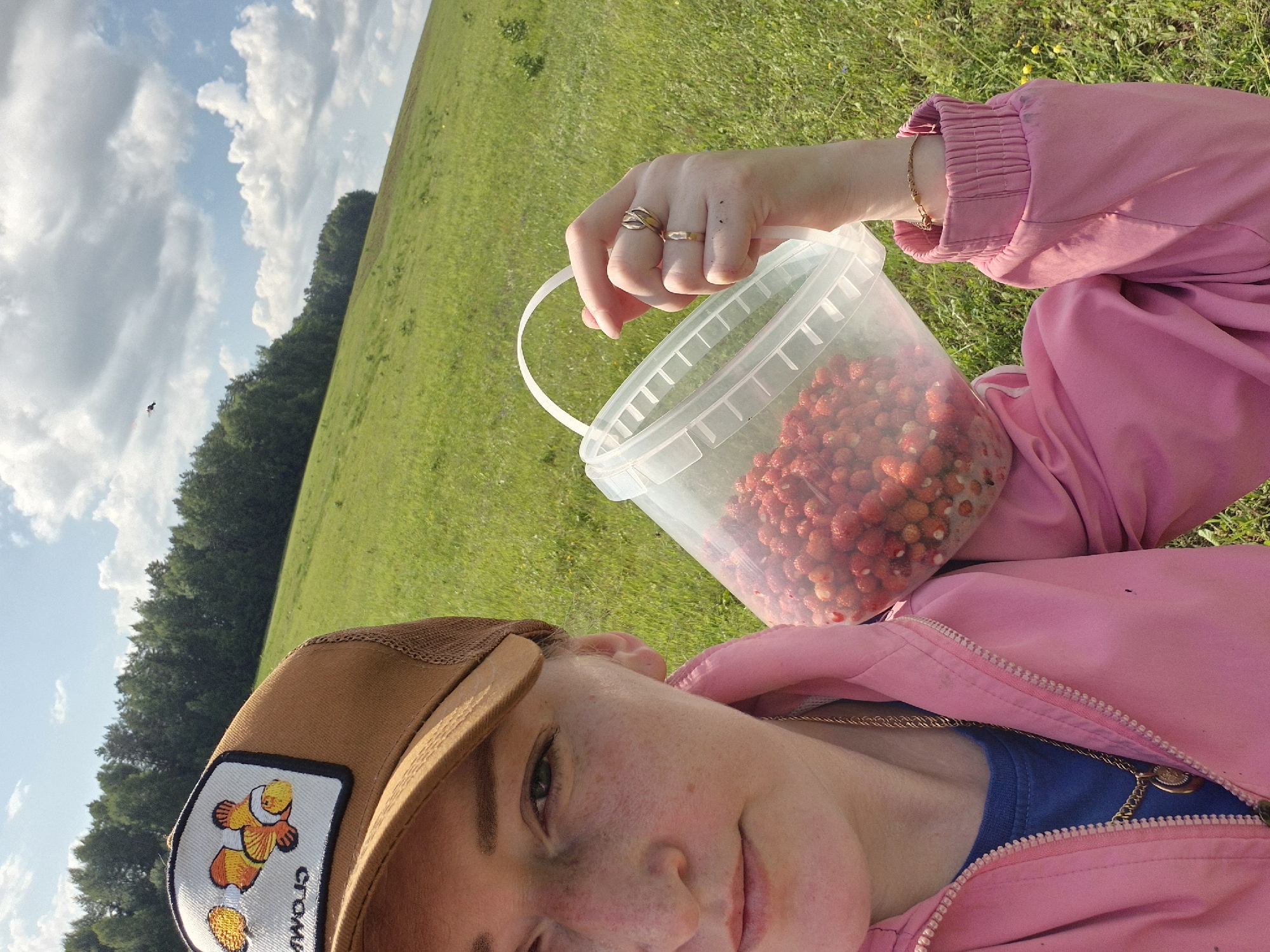 A woman wearing a pink jacket and a brown baseball cap with a clownfish patch stands in a grassy field under a partly cloudy sky. She is holding a clear plastic bucket filled with strawberries over her shoulder with her right hand. Her left hand is visible near her face. The background shows a sloping green hill with a line of trees at the top. A small bird is visible in the sky. The image appears to be a selfie taken outdoors.