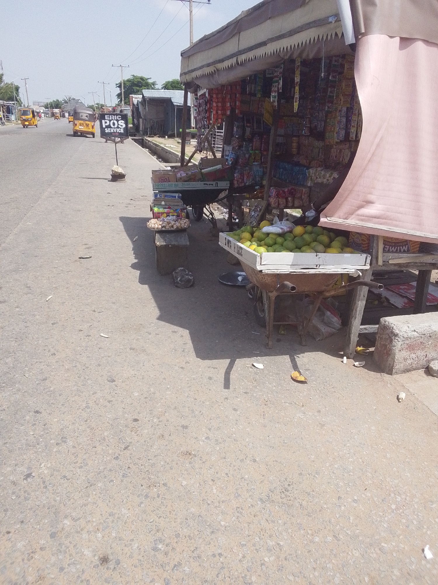 The image depicts a street scene in Jalingo, Nigeria, on a bright, clear day. The foreground is dominated by a paved road and sidewalk. On the right side of the image, a market stall is set up. The stall features a variety of goods, including packaged snacks, drinks, and a large display of green fruits, likely mangoes, in a wheelbarrow. A pink awning provides shade over the stall. A concrete block sits to the right of the stall.
In the background, the road stretches into the distance, with several yellow motorized tricycles, known as "keke napep," visible. One tricycle has "ERIC POS SERVICE" written on a black sign mounted on a pole in front of it. Further down the road, more tricycles and what appear to be small buildings or shacks line the street. Power lines and poles are also visible, indicating the presence of infrastructure.
The overall impression is one of a bustling, everyday street scene in a Nigerian town. The bright sunlight suggests it is likely daytime, possibly mid-morning or afternoon, with no indication of rain or other adverse weather conditions. The setting appears to be a roadside market, typical of many urban and semi-urban areas in Nigeria. The presence of the market stall and the tricycles points to local commerce and transportation. No people are clearly visible in the image.