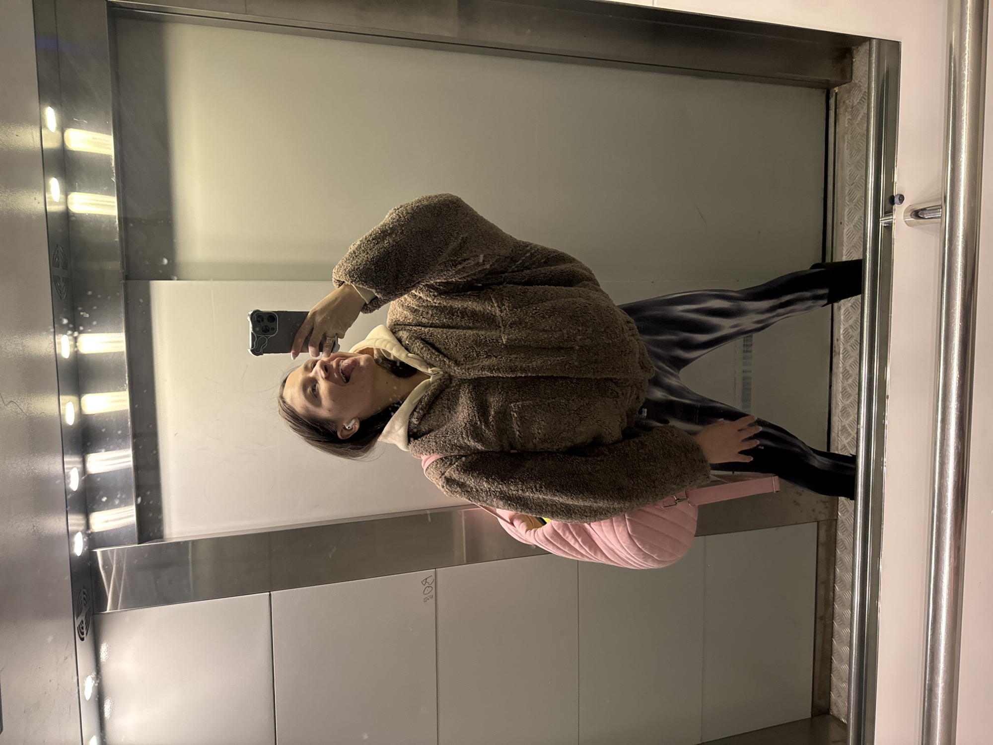 An indoor, vertical full-body mirror selfie shows a young woman inside what appears to be an elevator in Odintsovo, Russia. The woman, seen in reflection, is standing upright with her right arm raised, holding a dark smartphone with a light-colored case to take the picture. She is wearing a brown, fluffy, teddy bear-style jacket over a lighter-colored hooded top, and dark patterned leggings paired with dark shoes or boots. White wireless earbuds are visible in her ears. A pink quilted crossbody bag or fanny pack hangs on her left hip.
The setting is characterized by highly reflective walls, likely mirrors or polished metal, reflecting vertical strip lights on the left side of the frame. Metal trim surrounds the reflective panels, and a metallic diamond plate pattern is visible along the bottom edge where the wall meets the floor. A silver-colored metal handrail is present on the right side of the elevator interior. The artificial lighting illuminates the scene. The woman's mouth is slightly open as she looks towards her phone, capturing a casual moment. Faint, possibly handwritten numbers or letters, such as '208' or 'sos', are visible on a lower grey panel in the reflection.