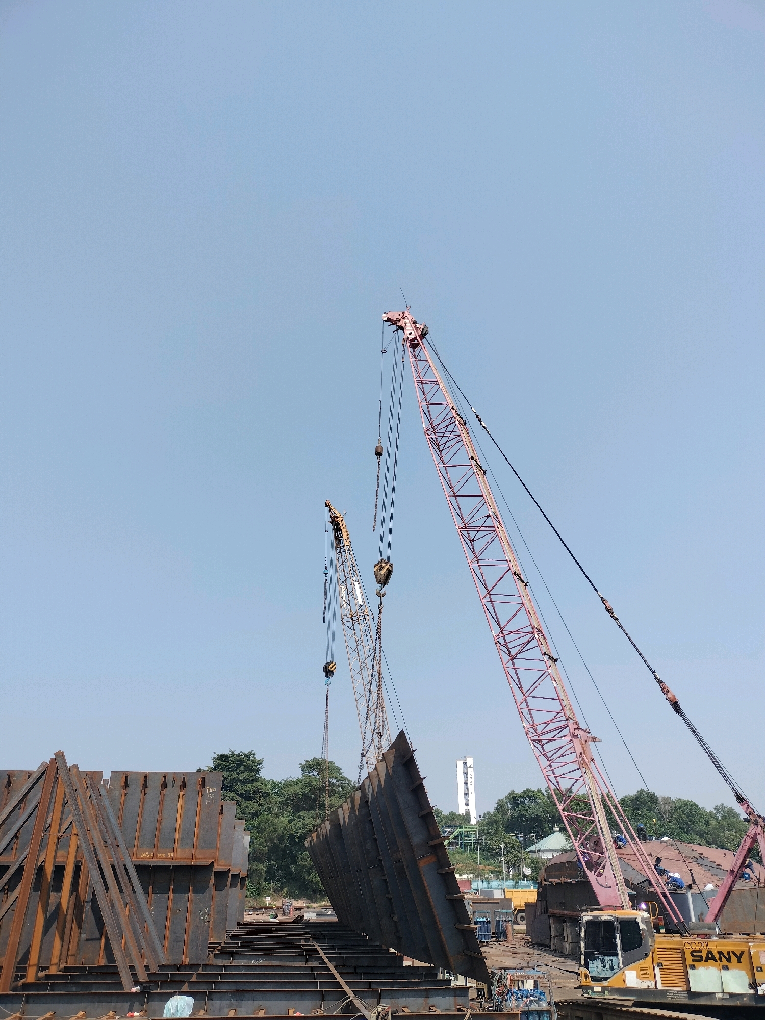 The image depicts a construction site in Batam, Indonesia, possibly within a shipyard or industrial area. Two large cranes are in operation: a tall, pink lattice boom crane on the right, and a smaller, yellow lattice boom crane on the left. The cranes are lifting and maneuvering large, dark metal sheets, likely part of a shipbuilding or construction project. In the foreground, a stack of similar metal panels is visible, leaning against each other. A yellow SANY excavator is parked in the lower right corner. In the background, there are trees, some buildings including a tall white tower with markings, and a light blue sky. Several workers, dressed in work clothes and hard hats, are visible on a structure in the middle ground, near the pink crane. The overall scene suggests active construction work is taking place.