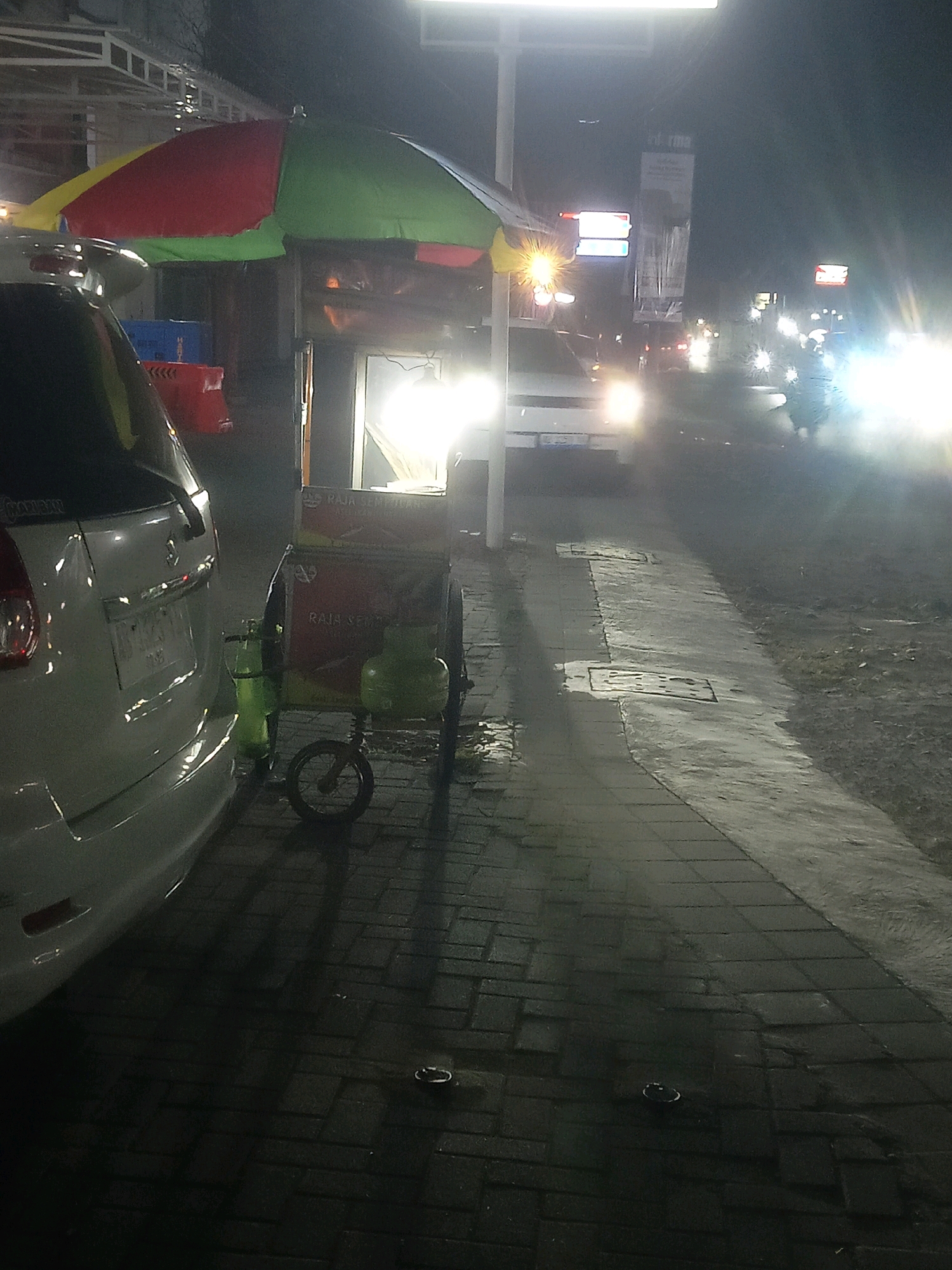 The image depicts a street scene at night in Trihanggo, Indonesia, likely near a roadside food stall. A white car is parked on the left side of the frame, partially obscuring a colorful umbrella covering a food cart. The food cart has a visible sign with red and yellow lettering that appears to say "RAJA SEMPURNA". Two green propane tanks are attached to the cart, along with a small wheel. The street pavement is made of dark bricks, and the area appears wet, suggesting recent rain. In the background, there are other vehicles, including motorcycles, with their headlights on, and illuminated signs. The overall lighting suggests it is nighttime, with artificial light sources from streetlights, vehicles, and the food stall itself. There are no visible people in the immediate foreground.