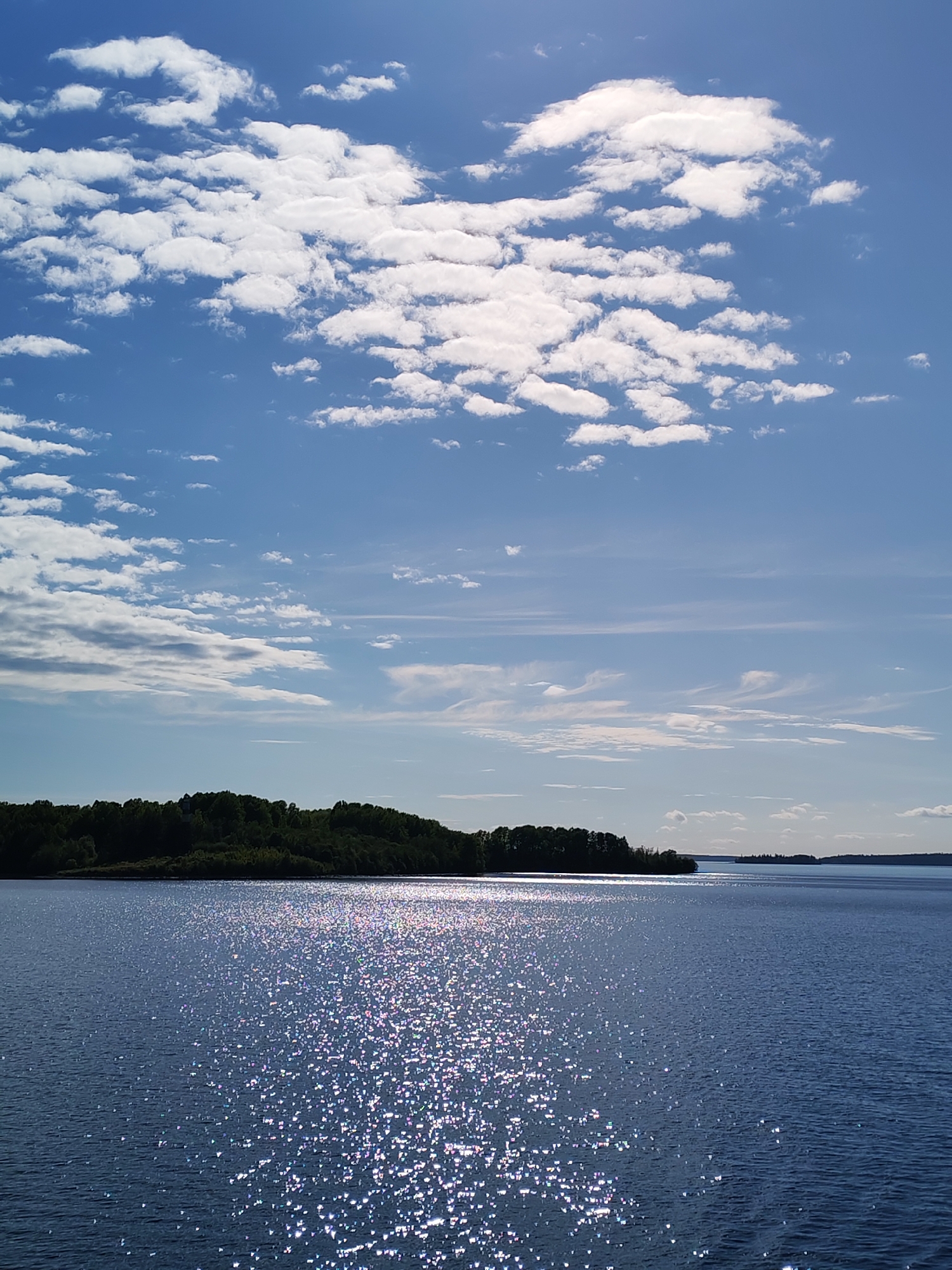 The image displays a serene natural landscape featuring a large body of water under a bright, partly cloudy sky. The sky, occupying roughly two-thirds of the frame, is a vibrant blue with numerous white clouds. These clouds vary in form, with some appearing as fluffy cumulus masses and others as more elongated, wispy cirrus streaks, particularly visible higher up and towards the horizon. The sun, though not directly in the frame, is positioned to reflect intensely off the water's surface, creating a dazzling pattern of shimmering white and iridescent sparkles that stretch from the foreground into the mid-distance. This brilliant reflection suggests a sunny midday or early afternoon.
A dark, densely forested landmass or island forms the distant shoreline, silhouetted against the brighter sky and water. It appears as a continuous line of green trees, providing a clear horizon line. Further to the right in the distance, another faint landmass is visible, indicating the extensive breadth of the water body, likely a large lake or wide river, consistent with the geographical features found in regions like Krivonogovskaya, Russia. The water itself is a deep blue-indigo hue, appearing calm with gentle ripples.
No people or explicit activities are visible, emphasizing the untouched natural beauty of the scene. The weather is clear and bright, contributing to the tranquil and peaceful atmosphere of the landscape. There is no visible text in the image.