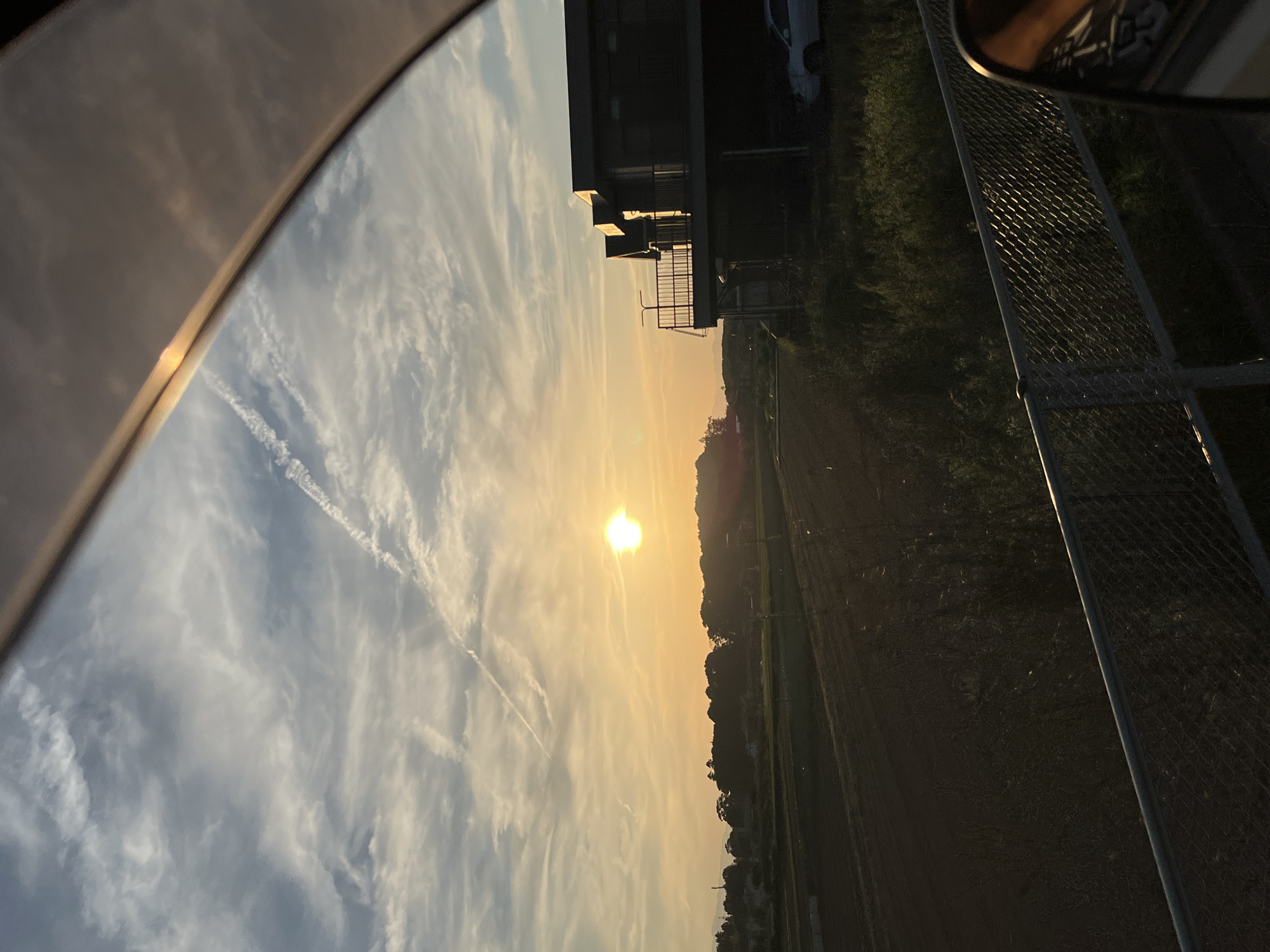 The image captures a sunset scene, viewed through a vehicle's window, possibly a car. The sky is a gradient of blue and orange, with streaky clouds and contrails visible. The sun is positioned near the horizon, appearing as a bright orb.
On the right side of the image, there is a building with a visible balcony and a car parked nearby. A treeline borders the building and extends towards the horizon. In the foreground, a portion of the vehicle's interior is visible.
The location is characterized by rural surroundings and a lack of distinguishing urban features, indicating it is likely outside a major city center. There is no way to know location based on this information. No people are directly visible in the scene.