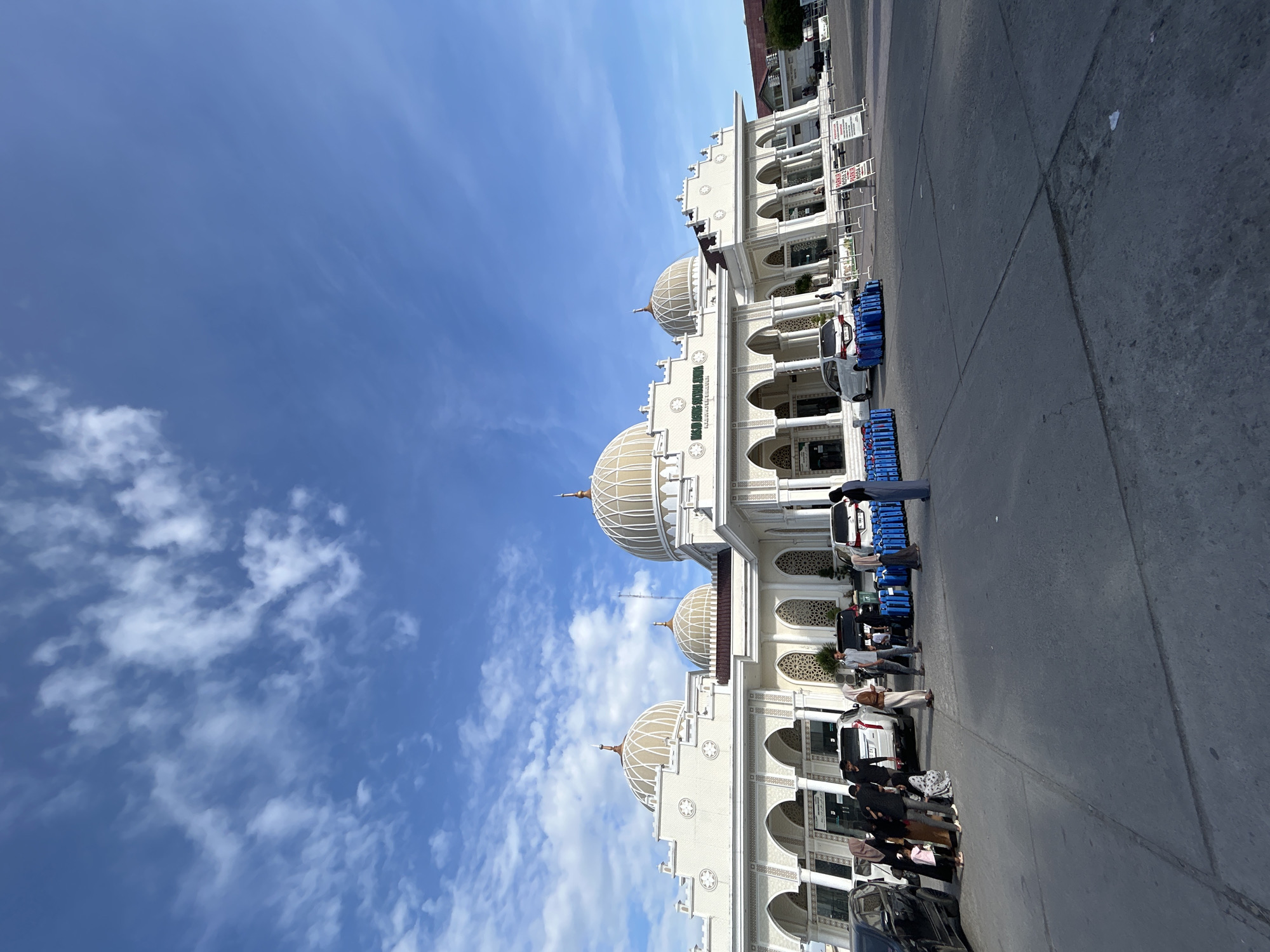 The image shows the exterior of the Masjid Agung Sultan Jeumpa, a mosque located in the city of Bireuen, Indonesia. The mosque features a grand white facade with multiple domes and arched entrances. Several cars are parked in front of the mosque, and a group of people are gathered in the foreground, some with luggage. The sky is blue with scattered clouds. The scene appears to be during the daytime. Signs are visible in front of the building, some indicating parking regulations. The overall atmosphere suggests a public space, potentially a transit point or a place of gathering.