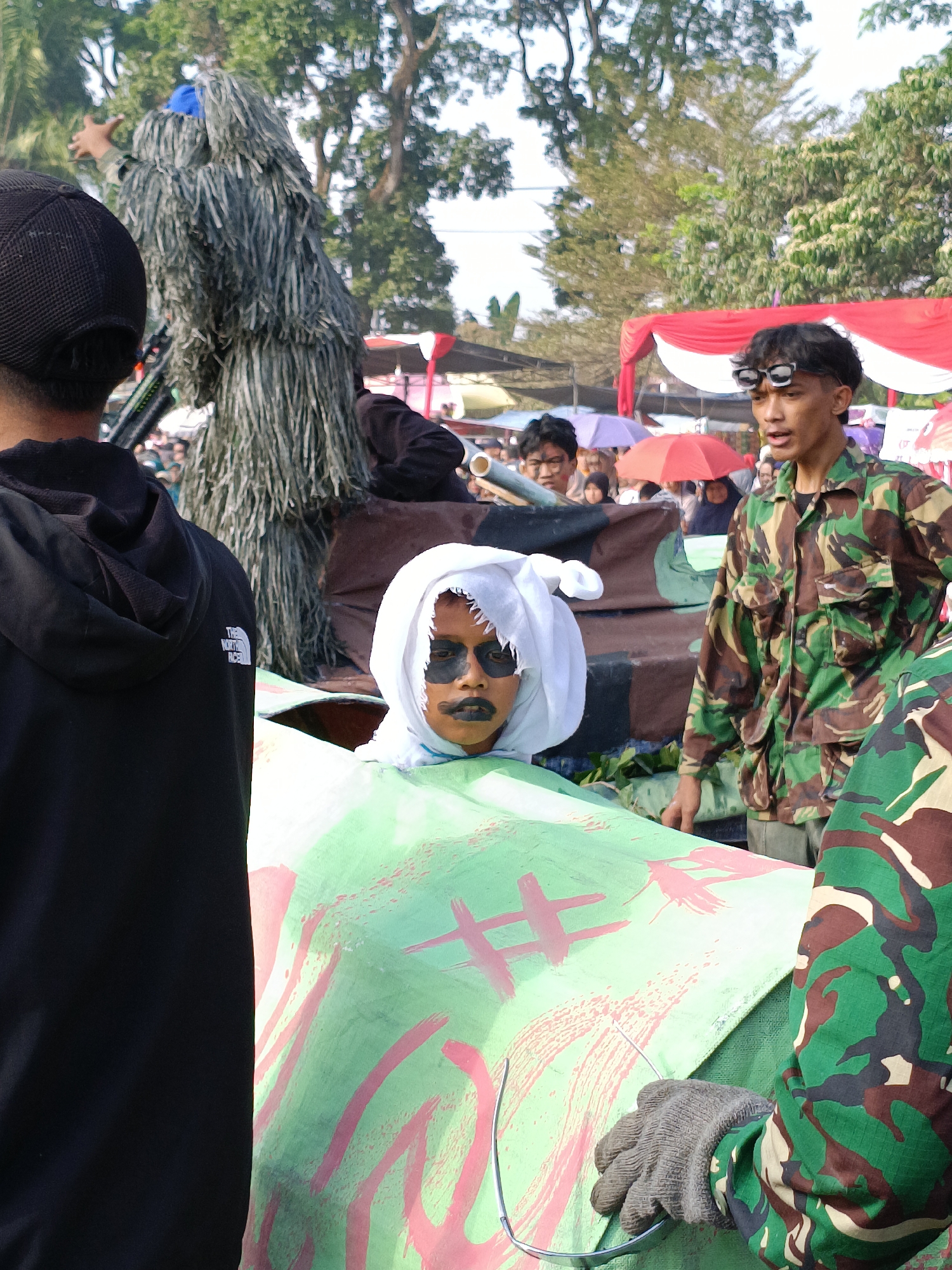 In Cibodas, Indonesia, a street scene unfolds with several individuals participating in what appears to be a festive or ceremonial event, possibly a parade. In the foreground, the back of a person wearing a black hoodie is visible. To their right, a child is positioned inside a large, green object adorned with abstract red markings and a white head covering. The child's face is painted with dramatic black markings around the eyes and mouth.
Further to the right, a young man in a camouflage uniform, complete with goggles, stands alert. Behind him, another individual in a ghillie suit, designed for camouflage with its shaggy, leaf-like material, is a prominent figure. The background reveals a gathering of people, some holding umbrellas, and tents with red and white banners, suggesting a public event or market. Trees with lush green foliage form the backdrop under a bright sky. The overall atmosphere suggests a lively community gathering.