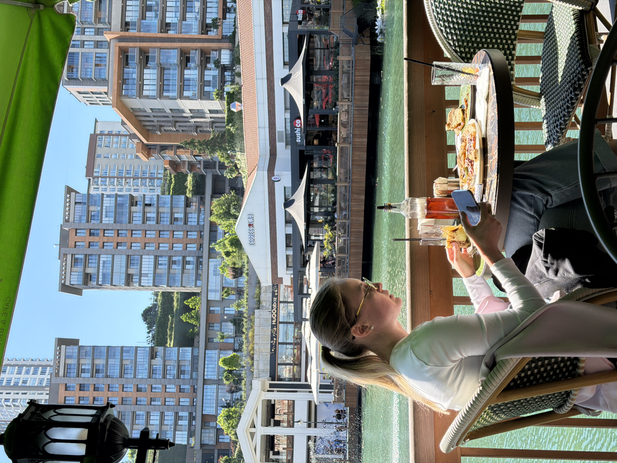 A vertical outdoor shot shows a woman seated at a table near a waterfront in Küçükçekmece, Turkey. The location appears to be a restaurant or cafe.
The woman has blonde hair in a ponytail and is wearing glasses, a white long-sleeved shirt, and a pink undergarment. She is holding a smartphone in one hand and a piece of food in the other. A table next to her has a plate of food, a bottle of liquid, and a glass with a straw.
The environment includes an artificial waterway and modern buildings in the background. Businesses with storefronts are visible, including "sushi co" and "ESPRESSOLAB," with the name "HUQqaBaz" printed near the latter. A lamp post stands near the left edge of the frame.