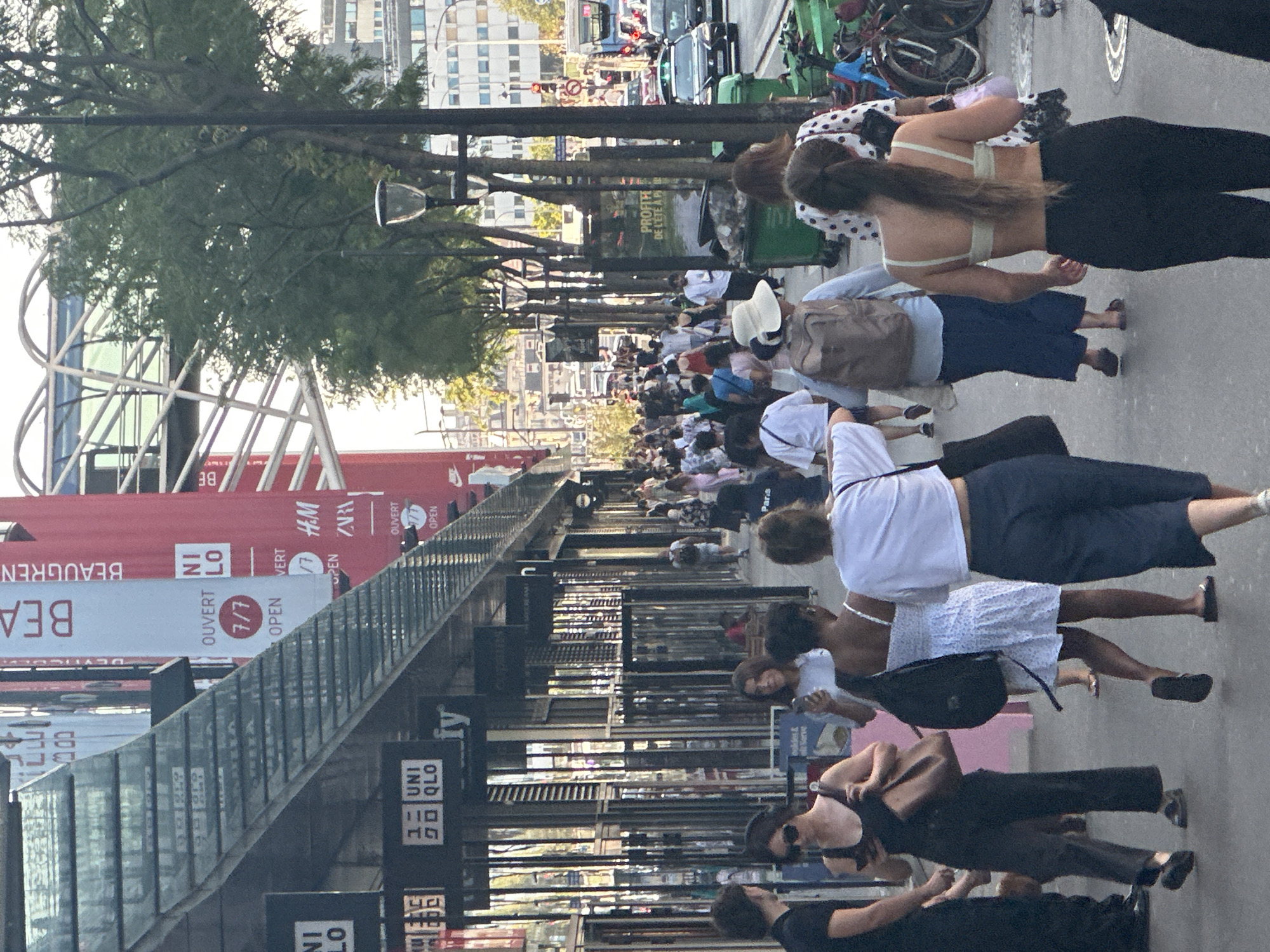 The image depicts a bustling street scene in Paris, France, likely along a commercial thoroughfare, judging by the prominent storefronts and signage. The perspective is from ground level, looking down the sidewalk and street.
On the left side of the image, there is a row of shops with large glass windows and awnings. Signs for "UNI QLO" are visible, indicating retail establishments. Above the shops, a modern, metallic structure with glass elements spans overhead. Red banners with white text, including "BE" and "OUVERT 7/7" (Open 7/7), are displayed vertically, suggesting businesses are open daily. Further down the street, trees line the sidewalk, with lampposts interspersed among them.
A diverse group of people are walking along the sidewalk, some in small groups and others individually. They are dressed in casual summer attire, including dresses, t-shirts, and pants. Several individuals are carrying bags or backpacks. Some are looking at their phones. The street itself has traffic, with cars and a bus visible in the background. Green garbage bins are positioned along the sidewalk. The overall atmosphere suggests a vibrant urban environment on a daytime.