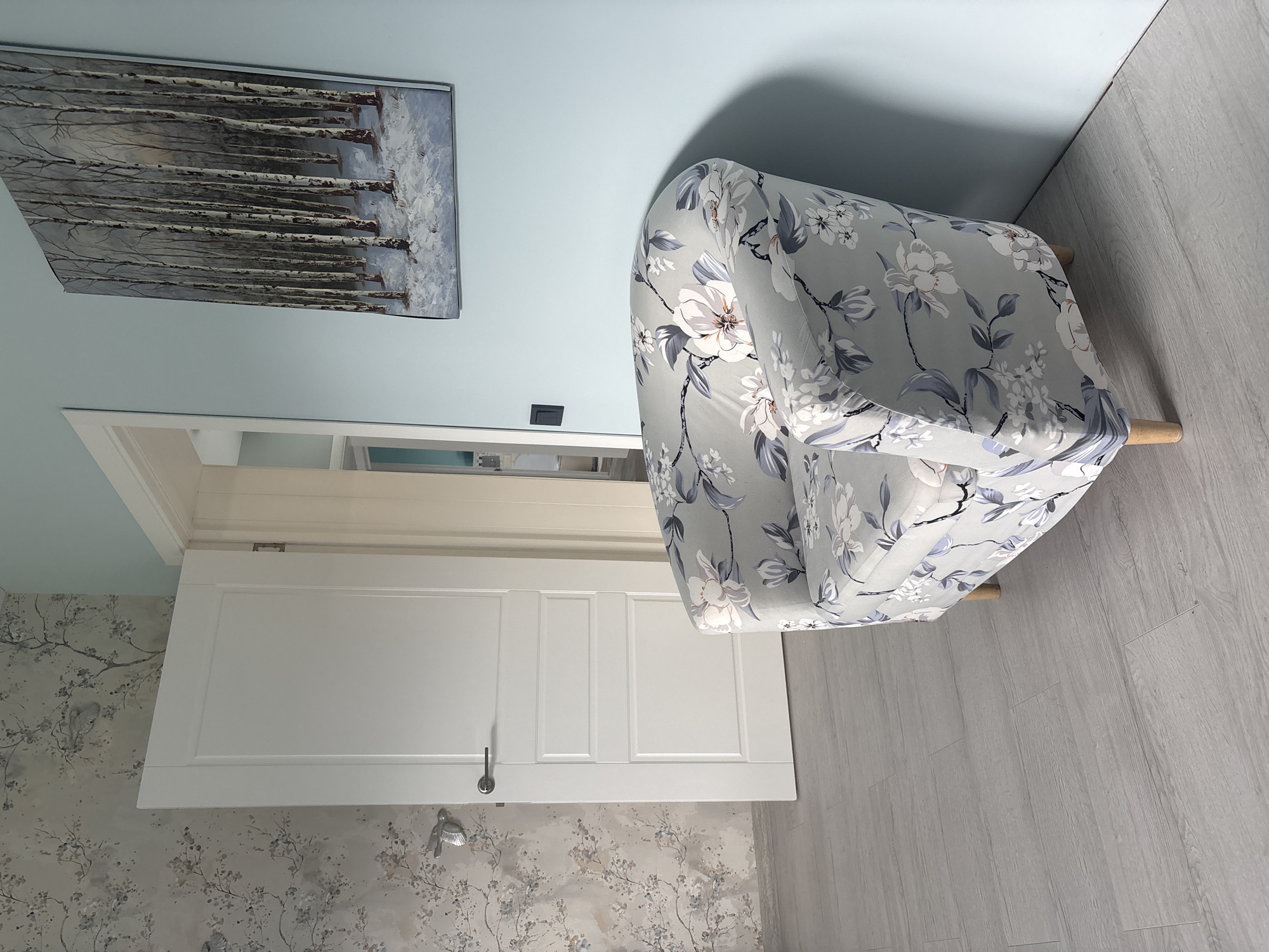 The image shows an interior scene. In the foreground, there is a floral armchair with a grey background and white and blue flowers. The armchair has wooden legs and is placed on a light grey wooden floor. To the left of the armchair is a white interior door with a dark handle. The wall to the left of the door is covered with floral wallpaper. To the right of the armchair, there is a light blue wall with a framed painting of a forest in winter. A black light switch is visible on the blue wall. The scene appears to be within a residential or commercial space in Дольче Вита Кп, Russia.