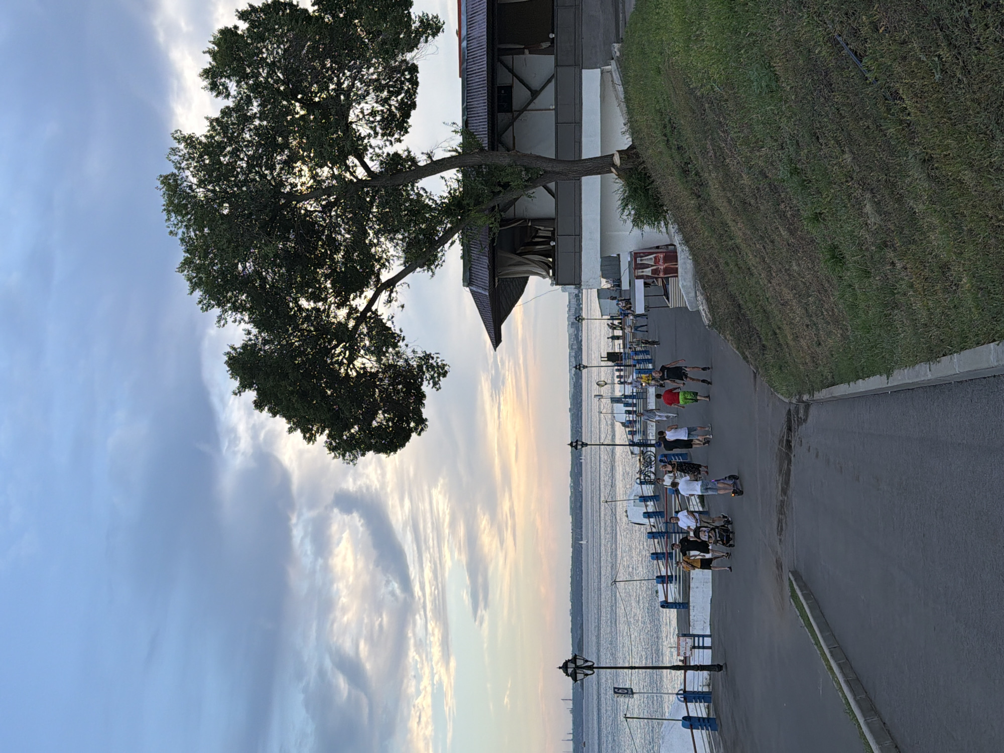 The image depicts a waterfront promenade in Saratov, Russia, during twilight. The sky is a mix of blue, gray, and soft pink hues, with scattered clouds. A large, mature tree with a dark green canopy dominates the right side of the frame, partially obscuring a building with a dark, corrugated roof and white lower structure.
Along the promenade, several people are visible. Some are walking, while others are pushing a stroller or riding a scooter. A group of individuals in casual attire are strolling near the water's edge, where a blue and white railing separates the walkway from the water. Two lampposts with traditional designs are spaced along the promenade. In the background, the broad expanse of a river or large body of water stretches towards a distant shoreline with industrial structures. The ground of the promenade is paved with asphalt, and a grassy, inclined embankment is visible on the right. Two Coca-Cola branded vending machines are positioned near the building. The overall atmosphere is serene, with people enjoying an evening walk by the water.