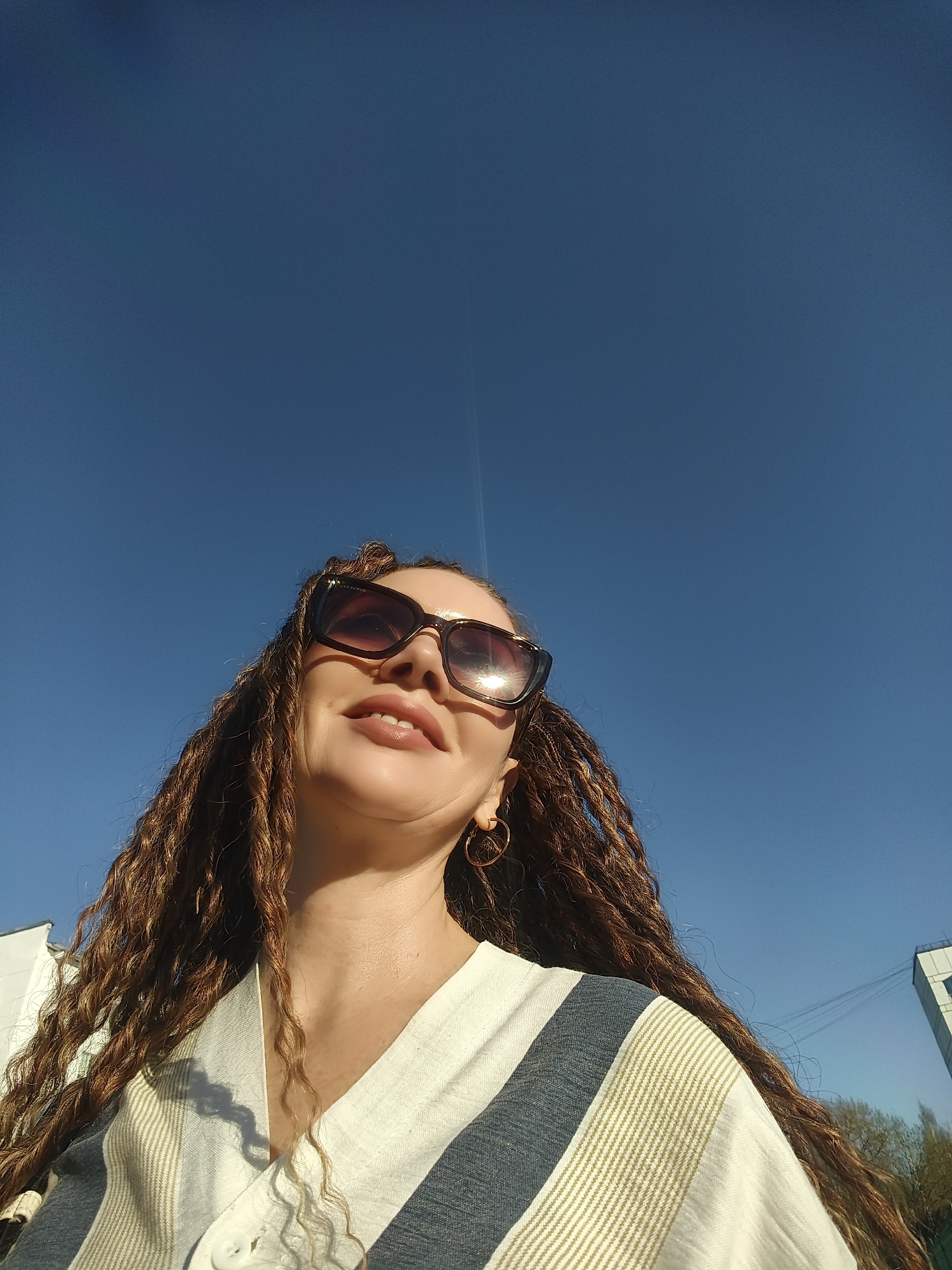 A low-angle shot features a woman with long, braided hair and sunglasses, set against a clear blue sky. She is wearing a light-colored shirt with vertical stripes. Her head is tilted upwards, and she is smiling slightly. The location is not identifiable from the image.