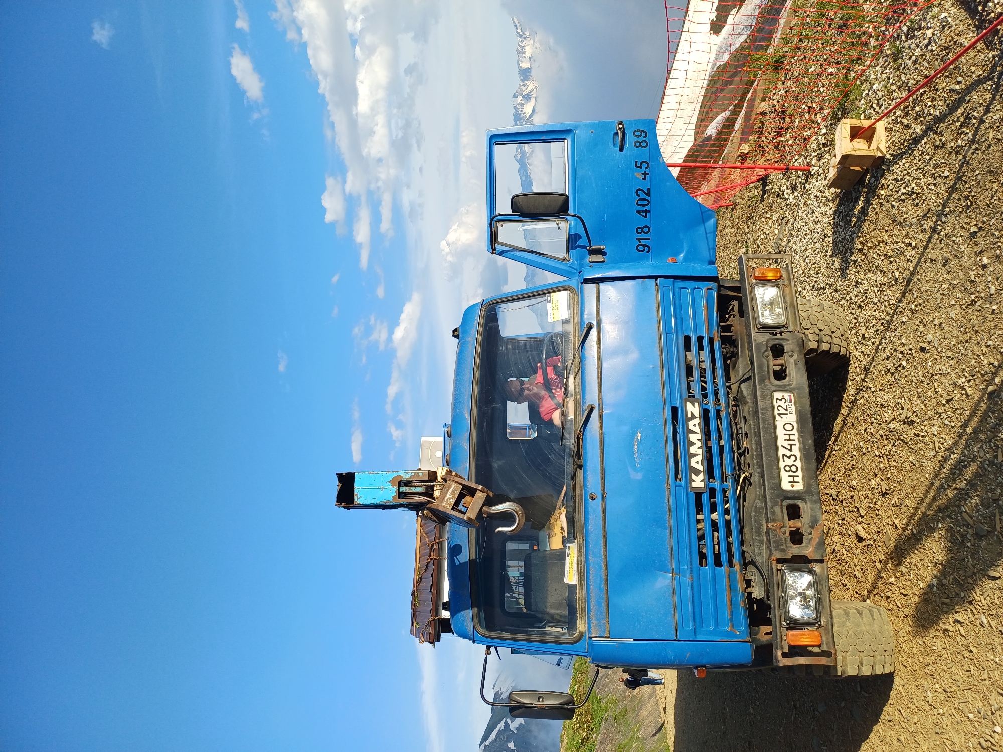 A blue KAMAZ truck with an open driver's side door is parked on a gravelly surface. A person wearing sunglasses and a pink shirt is sitting in the driver's seat. A mechanical crane with a hook is mounted on the back of the truck. In the background, there are snow-capped mountains, clouds, and a bright blue sky. A red safety net is visible to the right of the truck. The license plate on the truck reads "H834HO 123 RUS" and a phone number "918 402 45 89" is also visible on the side of the truck. Two people are standing in the distance to the left of the truck. The location is identified as Estosadok, Russia.