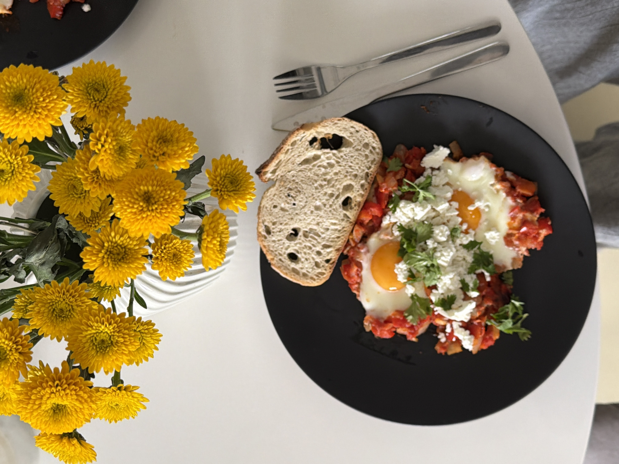 An overhead shot captures a breakfast or brunch setup on a white table, likely in an indoor setting in Khimki, Russia. The central subject is a black, round plate holding a serving of shakshuka. The dish features two fried eggs with vibrant orange yolks, surrounded by a rich, red tomato and pepper sauce, generously topped with crumbled white cheese (likely feta) and fresh green herbs, possibly cilantro or parsley. A thick slice of artisanal bread, showing an open, airy crumb structure, rests beside the plate.
Above the main plate, a silver fork and knife are neatly placed. To the left, a lively bouquet of bright yellow chrysanthemums, or similar round, compact flowers, adds a burst of color from a white, fluted vase. A portion of another black plate is visible in the upper left corner. On the right side of the frame, a soft grey textile is partially visible. The lighting is bright and even, suggesting daytime. The arrangement of food and flowers creates an inviting and pleasant atmosphere, implying a meal is either about to be enjoyed or has just been served. No text is visible in the image.
