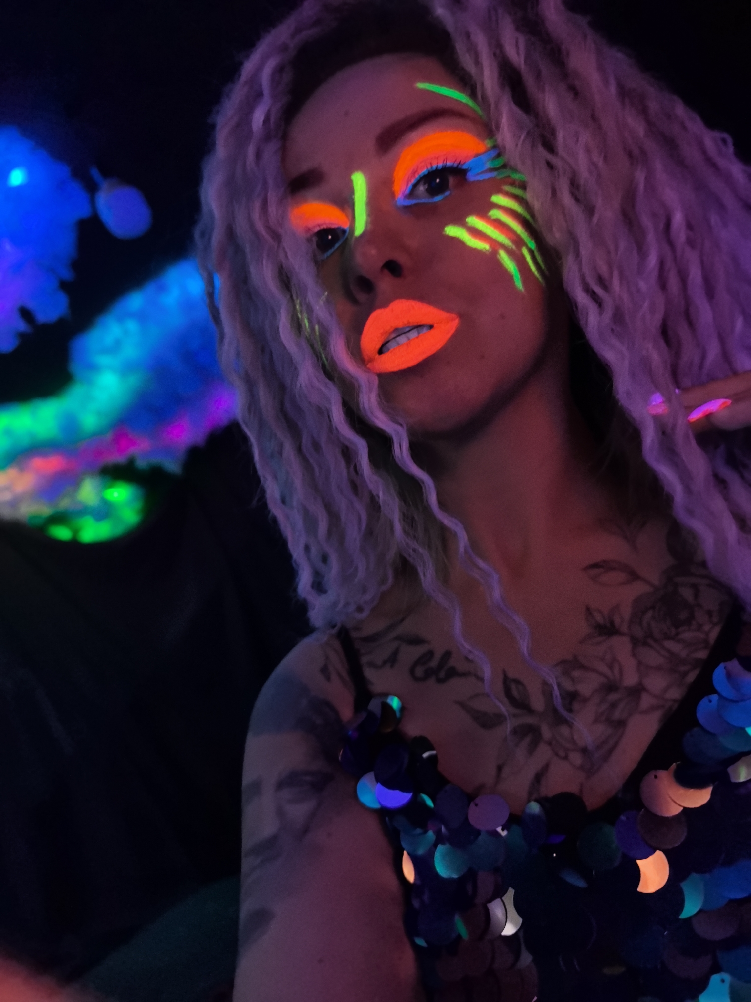 A vibrant portrait captures a woman in a dark, blacklight environment, adorned with striking, glow-in-the-dark makeup. Her face is accentuated with neon orange lipstick, a bold application of neon orange eyeshadow on her eyelids, and bright blue eyeliner along her lower lash line. Neon green stripes and dashes are painted on her cheekbones and near the inner corner of one eye. Her long, wavy hair appears light purple or lavender under the UV light. She wears a dark top embellished with iridescent sequins that reflect various neon colors, including blue, green, and orange. Visible tattoos include a large floral design and indistinct text on her chest, and a portrait on her upper arm. Her fingernails are also painted with a glowing polish. In the background, abstract, glowing shapes and swirls of light in shades of blue, green, and pink/purple suggest an artistic or party setting under blacklight conditions, likely at night. The subject's expression is confident and direct.