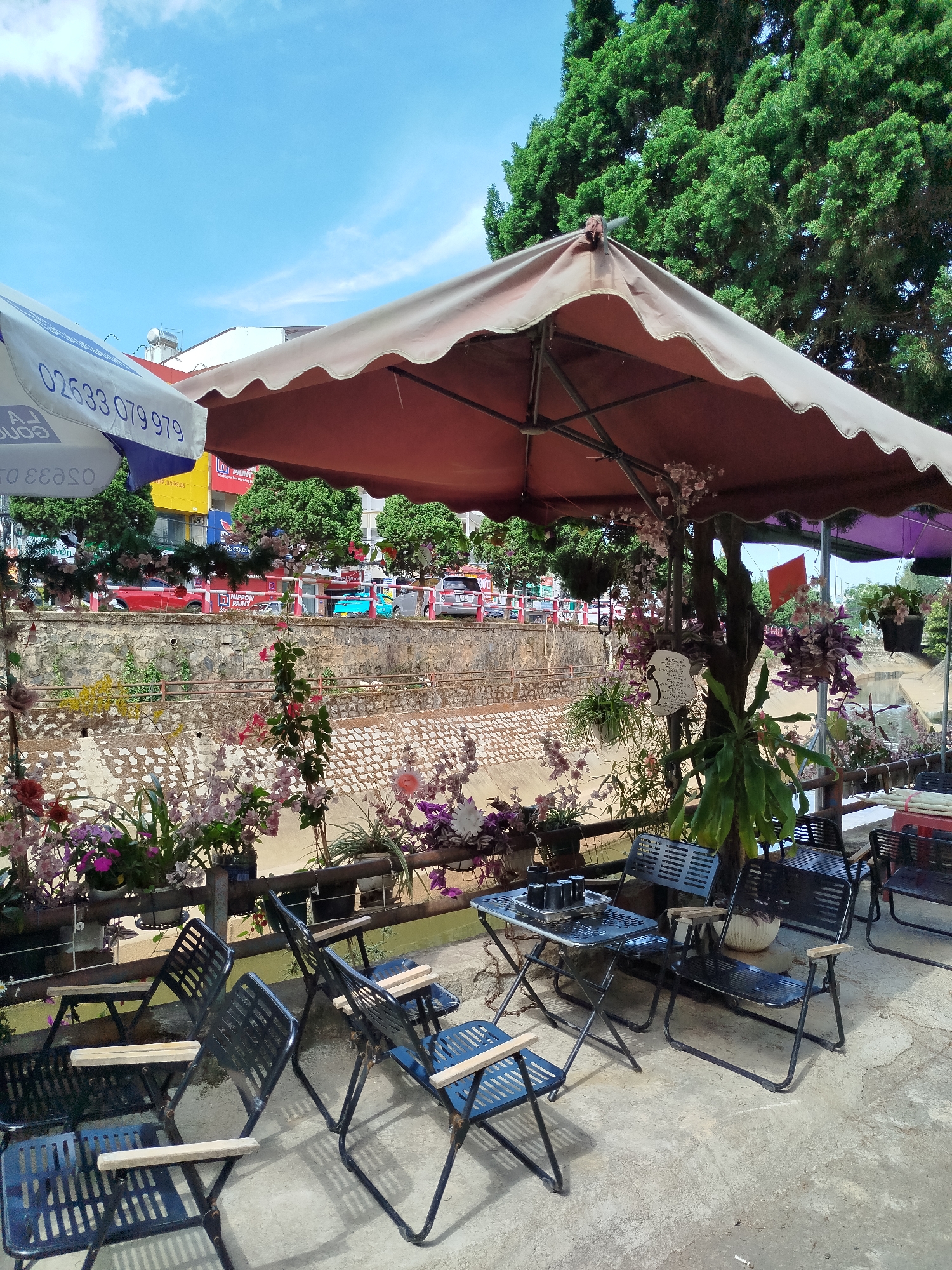 An outdoor seating area of a café in Da Lat, Vietnam, is depicted under a large brown umbrella. Several black metal chairs with wooden armrests are arranged around small tables. The seating area overlooks a waterway lined with a stone wall, featuring planters with various flowers and plants. Across the waterway is a street with buildings and trees. A white umbrella with blue text is visible on the left side of the image. The sky is blue with some clouds.