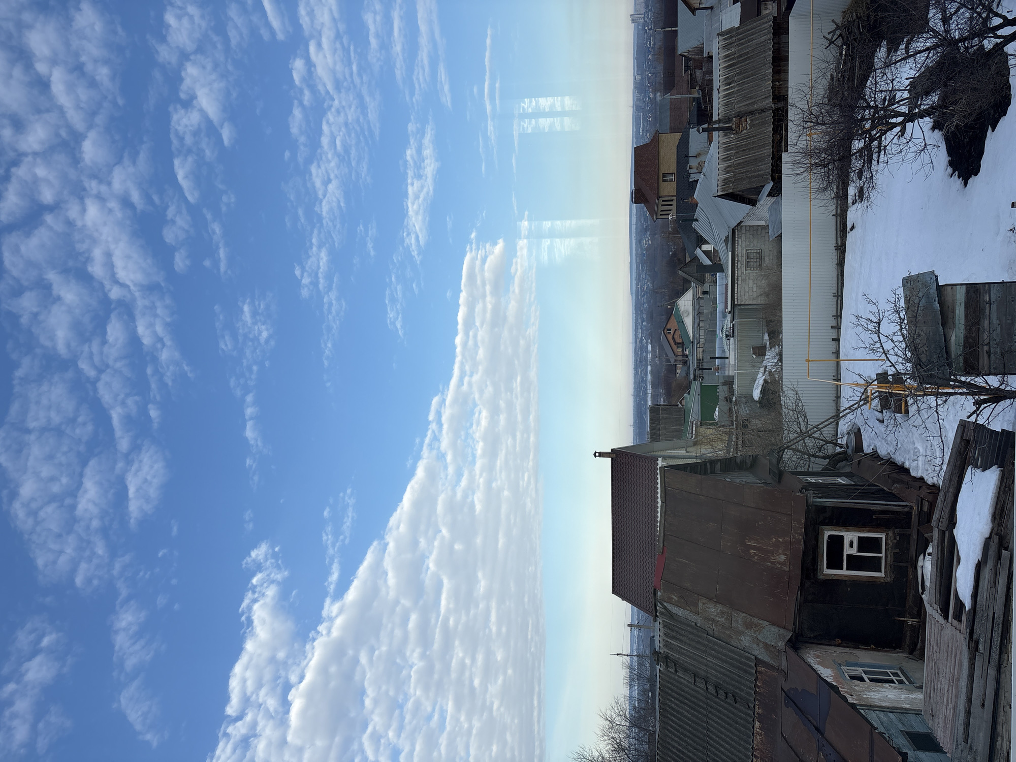 The scene depicts an elevated view over a residential area in Ulyanovsk, Russia, during a season characterized by patches of melting snow, suggesting late winter or early spring. The photograph was taken through a window, evidenced by faint reflections on the right side of the frame.
The upper half of the image features a bright blue sky partially covered by a scattered formation of altocumulus clouds, particularly dense along a horizontal band in the middle of the sky. The horizon line is hazy, revealing a distant cityscape with taller buildings.
The foreground and mid-ground comprise a densely packed cluster of single-story and two-story residential structures. Prominently in the left mid-ground is a weathered wooden house with a dark, possibly tarp-covered exterior and a single white-framed window. Its roof is made of rusty, corrugated metal. Adjacent to it, a section of a light blue-painted house with a similar roof is visible. Further back and slightly to the right, a more substantial two-story building with a red-tiled roof and a light-colored brick facade is discernible, featuring a chimney.
Property lines are demarcated by various fences, including a prominent light-colored corrugated metal fence running horizontally across the mid-ground. The ground between houses shows areas of exposed dark earth interspersed with irregular patches of melting snow. Bare deciduous trees and shrubs are scattered throughout the yards. A small, dark bird stands on the snow in a yard behind the corrugated fence in the central mid-ground. Utility poles and wires are visible above the rooftops, extending towards the distant horizon. No human figures are present.