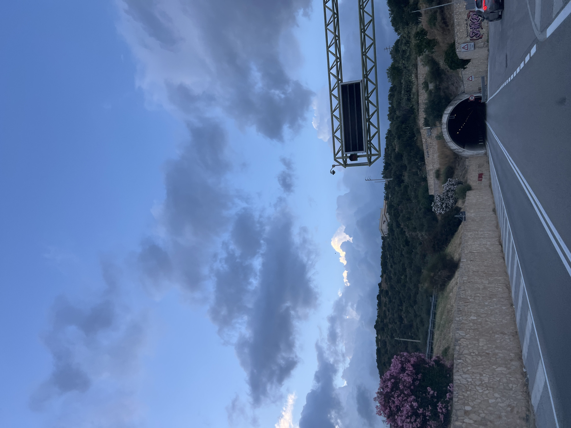 The image shows a view of a road leading towards a tunnel entrance in Mattinata, Italy. The sky is partly cloudy with a mix of dark and light gray clouds against a blue background, suggesting either early morning or late afternoon. On the left side of the road, there is a retaining wall constructed of rough, light-colored stones. Above the wall, a hillside is covered with dense green vegetation, with a large bush bearing pink flowers prominently in the foreground. Further up the hill, a building is partially visible.
The road itself is paved and marked with white lines, indicating two lanes of traffic. A large metal gantry with a dark rectangular display screen spans the road overhead, equipped with a camera on a pole attached to its structure.
The tunnel entrance is arched and illuminated from within. To the right of the tunnel entrance, there are various warning signs, including a circular sign with "50" inside, indicating a speed limit. Graffiti is visible on the walls surrounding the tunnel entrance. A dark-colored car is stopped near the right edge of the frame, just before the tunnel. The overall scene captures a moment on a road approaching a tunnel in a hilly, possibly coastal, environment, characteristic of the Gargano region where Mattinata is located.