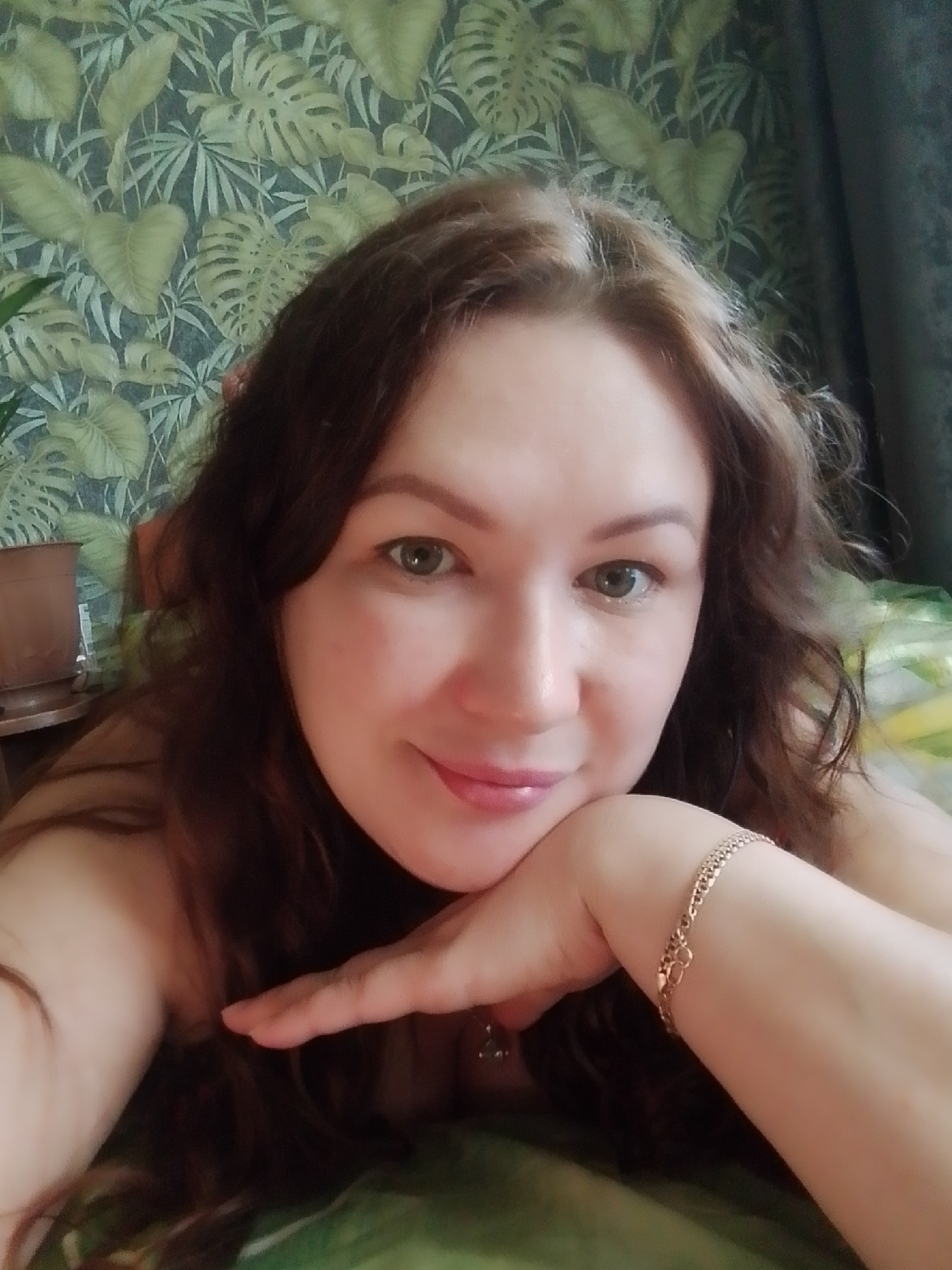 A woman with brown hair is resting her head on her hand, which has a gold bracelet on her wrist. She is fair-skinned and has light eyes. The background includes a wall with a leafy plant pattern and a houseplant in a terra cotta pot. The location is Noviy, Russia.