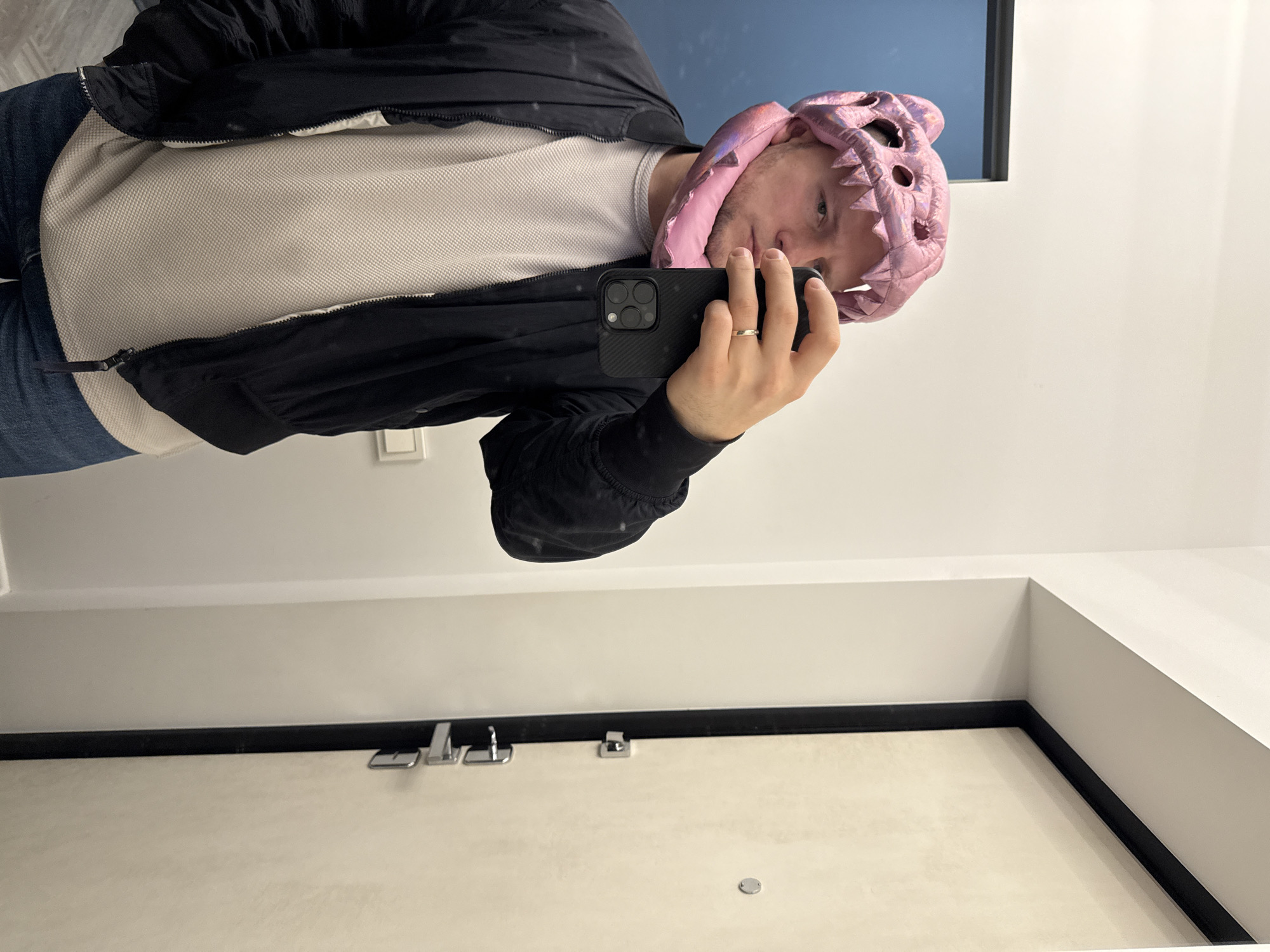 A person wearing a pink dinosaur-themed hat and a black jacket is taking a selfie in front of a light-colored door. The person is holding a black smartphone. The location cannot be determined without additional information about the specific building or surroundings visible in the image. It is not possible to confirm that the location is Krasnoyarsk, Russia, based on the provided image.