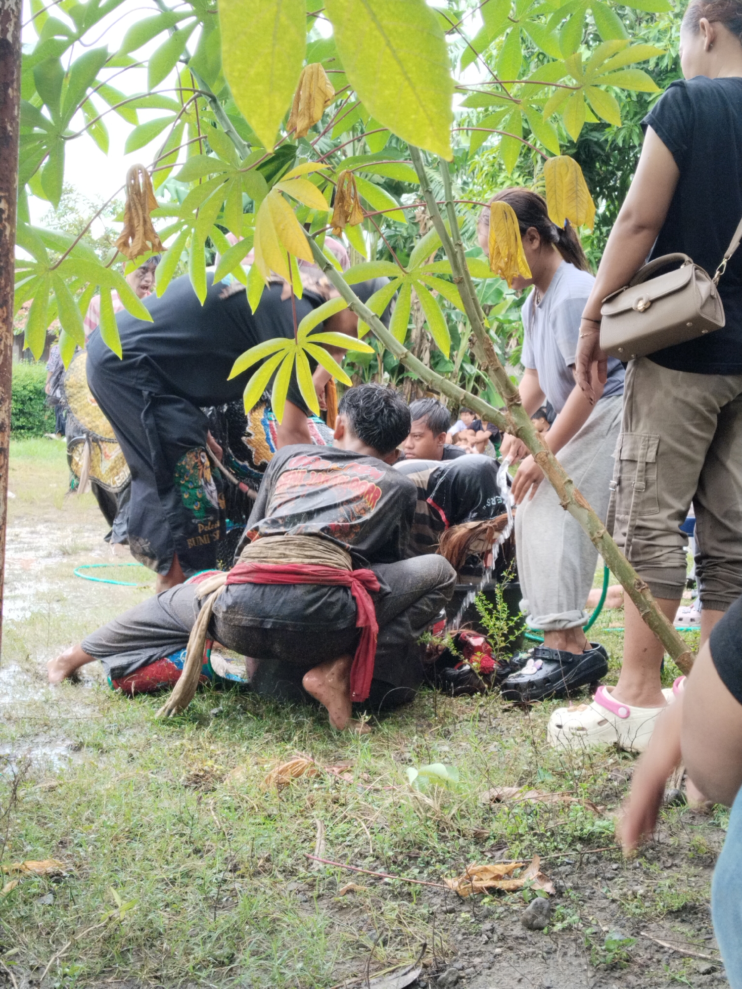 The image depicts a gathering of people in what appears to be an outdoor setting, possibly Karang Tengah, Indonesia, judging by the environment and cultural elements. There are individuals in traditional attire, some interacting closely near a small tree or plant. Water is being poured or splashed in the vicinity, indicating some sort of ritual or ceremonial activity. The ground is wet, suggesting recent rain or purposeful watering. Other people stand around observing the event, wearing more casual clothing. The scene appears to be a mix of performance and observation.