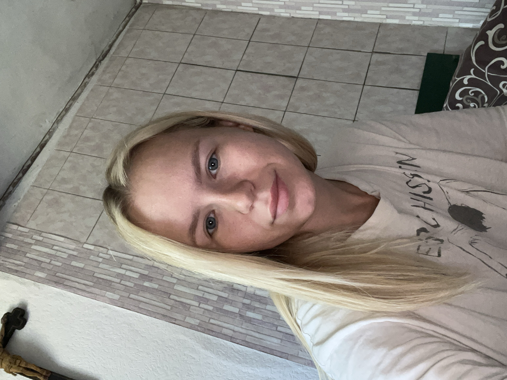The media file is a close-up, selfie-style photograph of a young woman with blonde hair. She is lying down, with her head tilted. Her eyes are open and she is looking towards the camera with a slight smile. She is wearing a light-colored t-shirt with some text printed on it, which appears to be in a dark, stylized font. The text is partially visible and reads "HISSIN".
The background is a tiled floor and wall, suggesting an indoor setting. There is a patterned pillow or cushion visible on the right side of the frame. A small green rectangular object is also visible on the floor. In the lower left corner, a dark object with a cross shape is partially visible, possibly a crucifix or religious icon. The lighting appears to be natural, suggesting it could be daytime. The overall mood of the image is relaxed and casual.