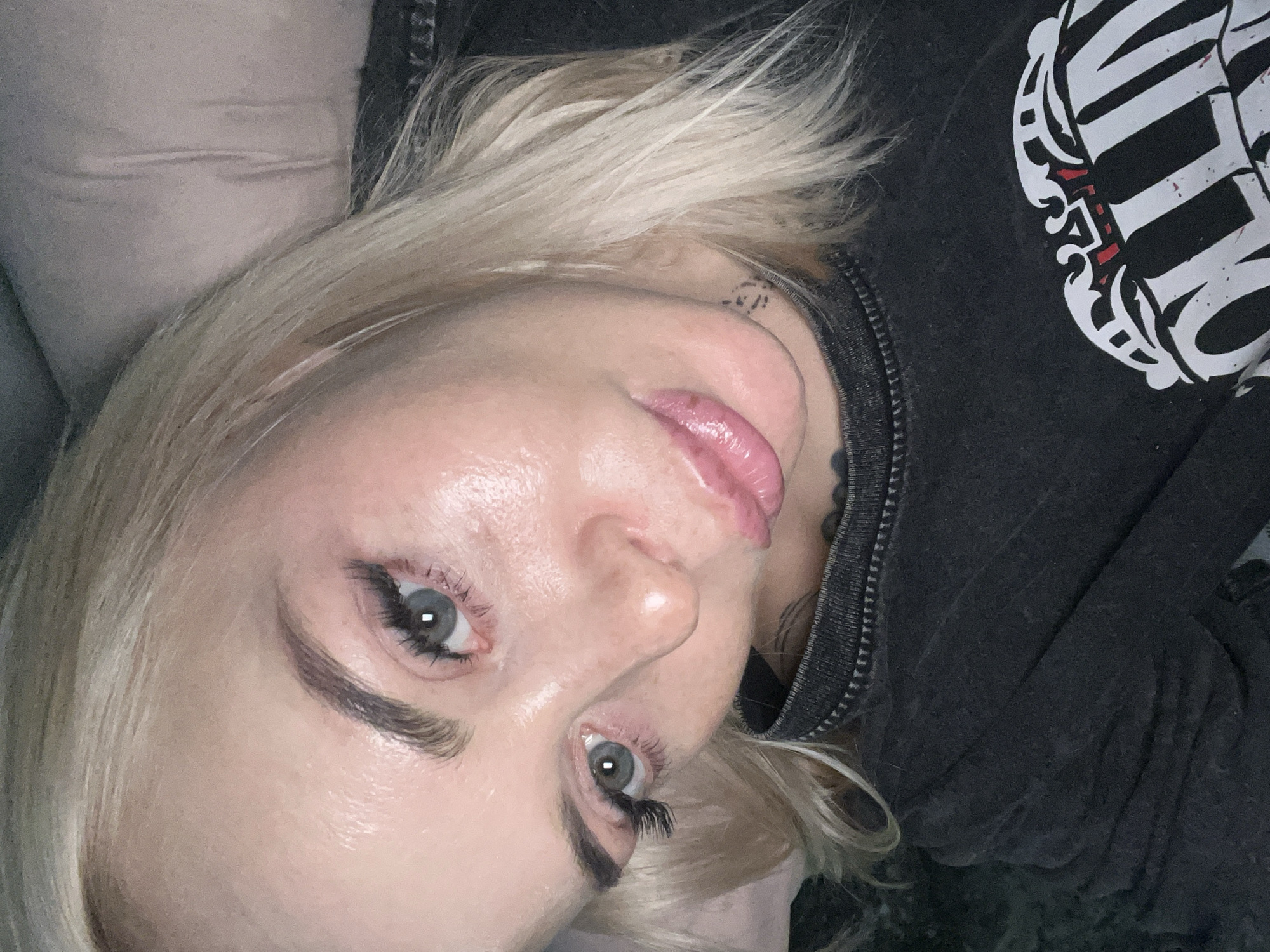 A close-up, vertically oriented portrait features a person, likely a woman, with fair skin and platinum blonde hair. She has striking blue-grey eyes, enhanced by dark, defined eyebrows and voluminous false eyelashes. Her lips are a glossy pink, slightly parted.
She is lying down, looking directly into the camera. A small, dark, abstract tattoo or symbol is visible on her neck, behind her right ear. She is wearing a dark, possibly charcoal grey, hooded sweatshirt or jacket, with a visible zipper detailing the neckline. The garment features a prominent white graphic design on the right side of her chest. This graphic appears to be stylized text or an emblem, possibly with red accents, and gives an edgy aesthetic. While partially obscured, elements resembling ornate 'W' or 'V' and other bold letters are visible.
The background is simple, consisting of a light, neutral-toned fabric, possibly a pillow or blanket, suggesting an indoor setting such as a bedroom or living room. The lighting is soft, highlighting her facial features. Her expression is neutral and direct, creating an engaging gaze.