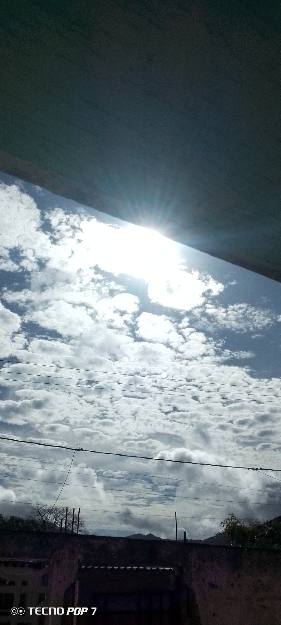 The image, taken with a TECNO POP 7 phone, shows a view of the sky in Altagracia, Venezuela. The sky is filled with white cumulus clouds, and the sun is shining brightly through a break in the clouds. Power lines are visible across the middle of the frame. In the lower portion of the image, there is a dark wall. The top of the image shows a dark, solid structure, potentially a roof or ceiling.