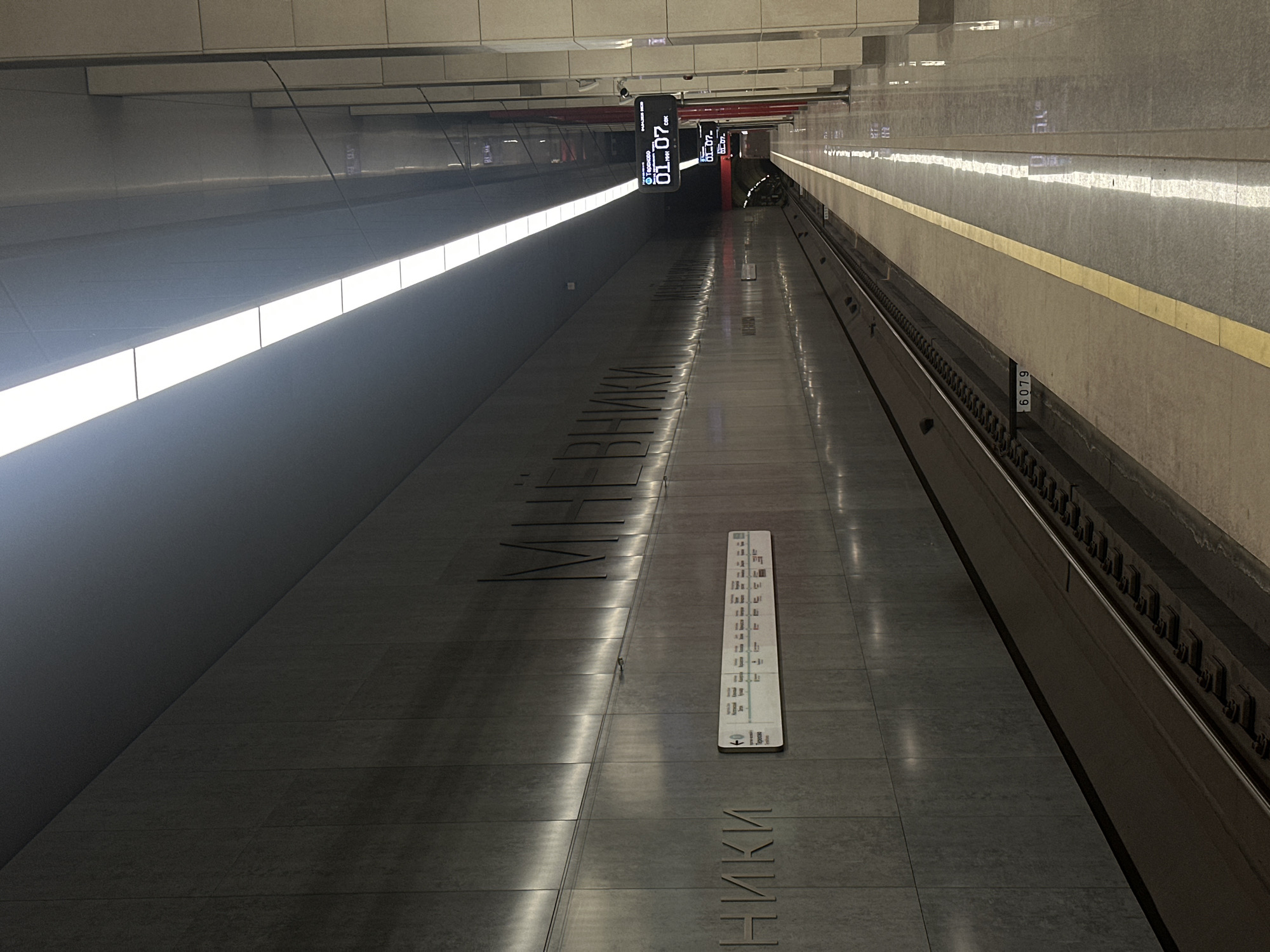 This is an image of the empty platform at the Chkalovskaya metro station on the Lyublinsko-Dmitrovskaya Line of the Moscow Metro, Moscow, Russia. The station features a gray color scheme and is well lit by ceiling-mounted fluorescent lights. A station map with Cyrillic text and an electronic display are visible. Tracks run along the right side.
