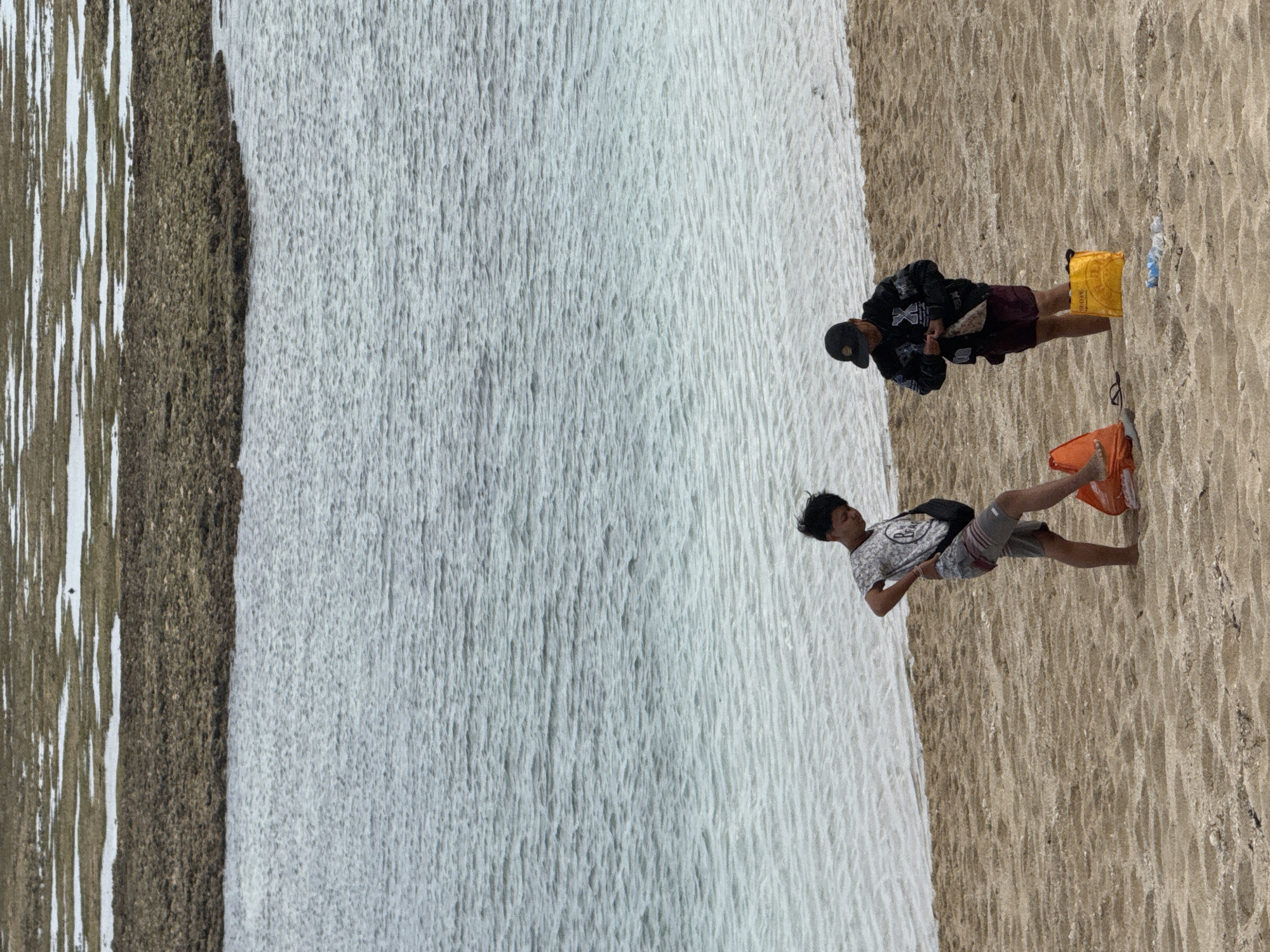 The image depicts a beach scene in Kuta Selatan, Indonesia. Two individuals are present on the sandy shore. The person on the left is a young male wearing a patterned t-shirt and patterned shorts, with a dark bag slung over his shoulder. He is standing with one leg extended forward, resting on an orange bag. The person on the right is wearing a dark jacket with a prominent white 'X' design, dark shorts, and a baseball cap. They are holding a small item in their hands. In the foreground, on the sand, are two bags: an orange one being used by the first individual and a bright yellow bag. Several plastic bottles are also scattered on the sand. The water in the background is a calm, clear blue, transitioning to a lighter turquoise near the shore, with visible ripples. Further in the background, there is a rocky, submerged area with patches of brown and white, suggesting shallow water or a reef. The sky appears overcast, contributing to the muted lighting of the scene.