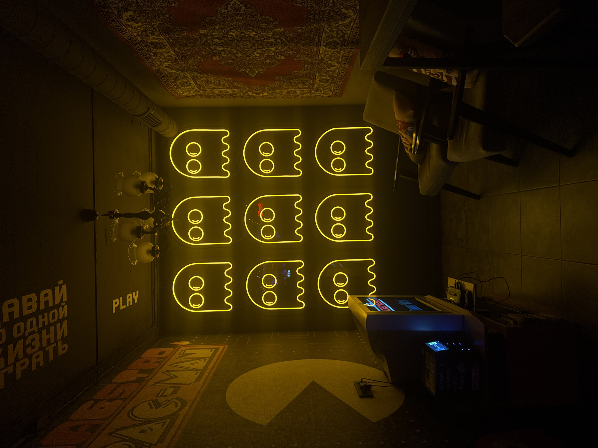 The image shows the interior of the Pac-Man-themed arcade bar “Pivoi!” in Hrodna, Belarus. The space is dimly lit with a yellow cast. A wall is adorned with nine neon light figures of Pac-Man ghosts arranged in a 3x3 grid. A vintage chandelier hangs to the left of the ghost figures. The word "PLAY" is emblazoned on the wall near the chandelier. A vintage, two-player Pac-Man arcade game stands to the right of the neon ghosts. In front of the game is a yellow Pac-Man figure design on the floor. The wall to the left of the chandelier features a phrase written in Cyrillic. A rug with an ornate pattern covers the ceiling. A chair and table are situated in the upper right corner of the image.