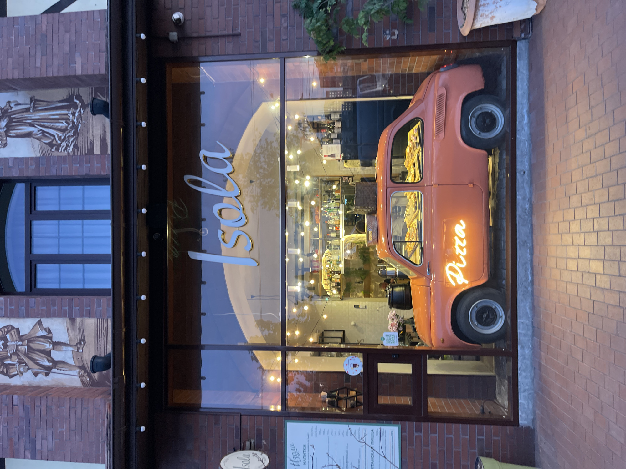 The image captures the exterior of the "Isola Pizza" restaurant in Kaliningrad, Russia. The restaurant's storefront features a large glass window displaying a vintage orange car with "Pizza" written on its side in neon lights. The interior of the restaurant is visible through the window, illuminated with string lights and showcasing a bar area stocked with bottles. A menu board and the restaurant's logo are positioned at the bottom of the window. The building's exterior includes brickwork and decorative wall art depicting historical figures.