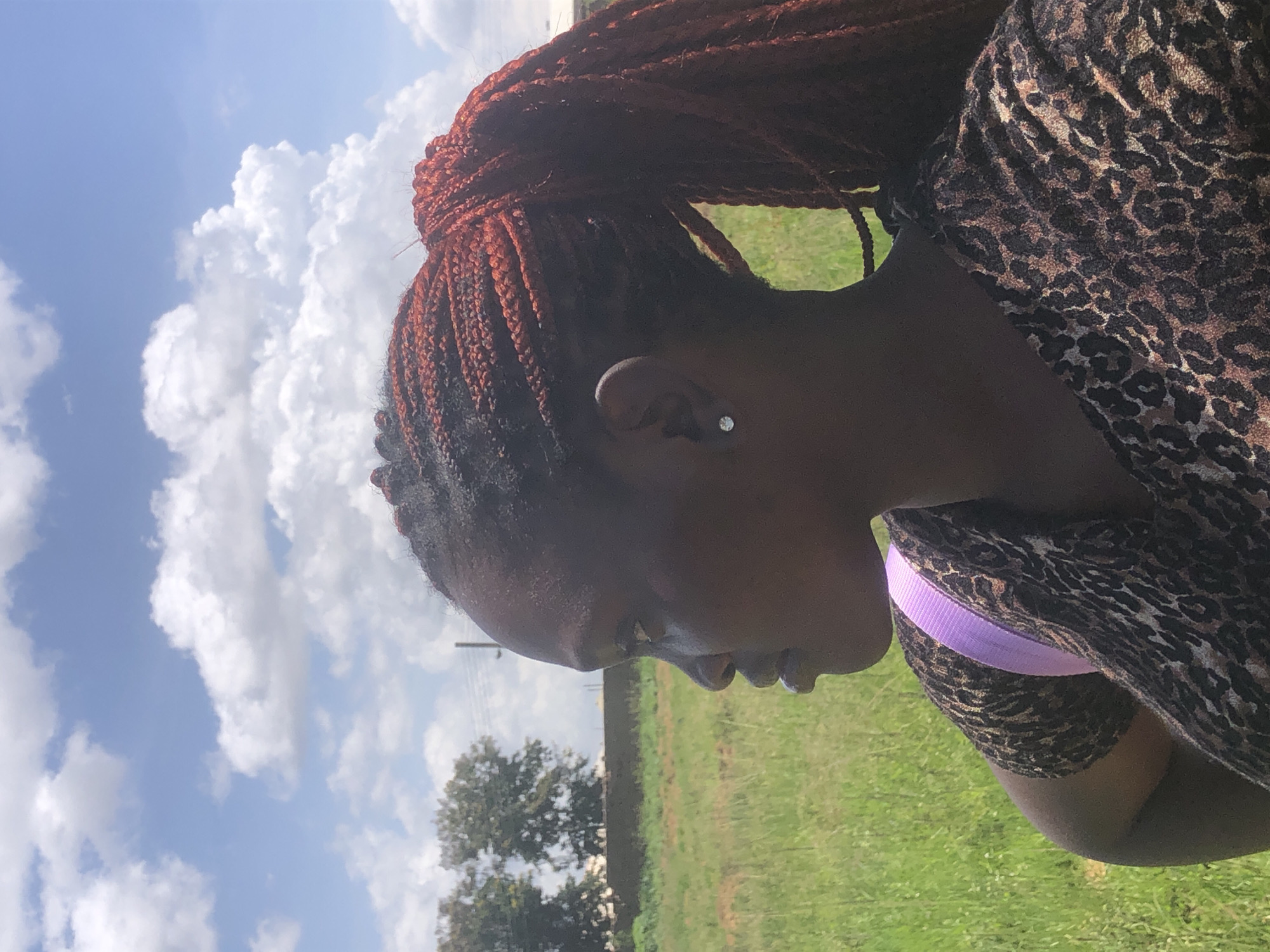 A close-up, profile view shows a dark-skinned woman with braided, reddish-brown hair pulled back into a ponytail. She is wearing a leopard-print top and a light purple strap crosses her shoulder. A small, clear stud earring is visible in her right ear. She is outdoors, with a grassy field and trees in the background under a partly cloudy blue sky. The location appears to be a rural or semi-rural area, potentially on the outskirts of Gwafan, Nigeria, given the natural landscape. The overall lighting suggests it is daytime.