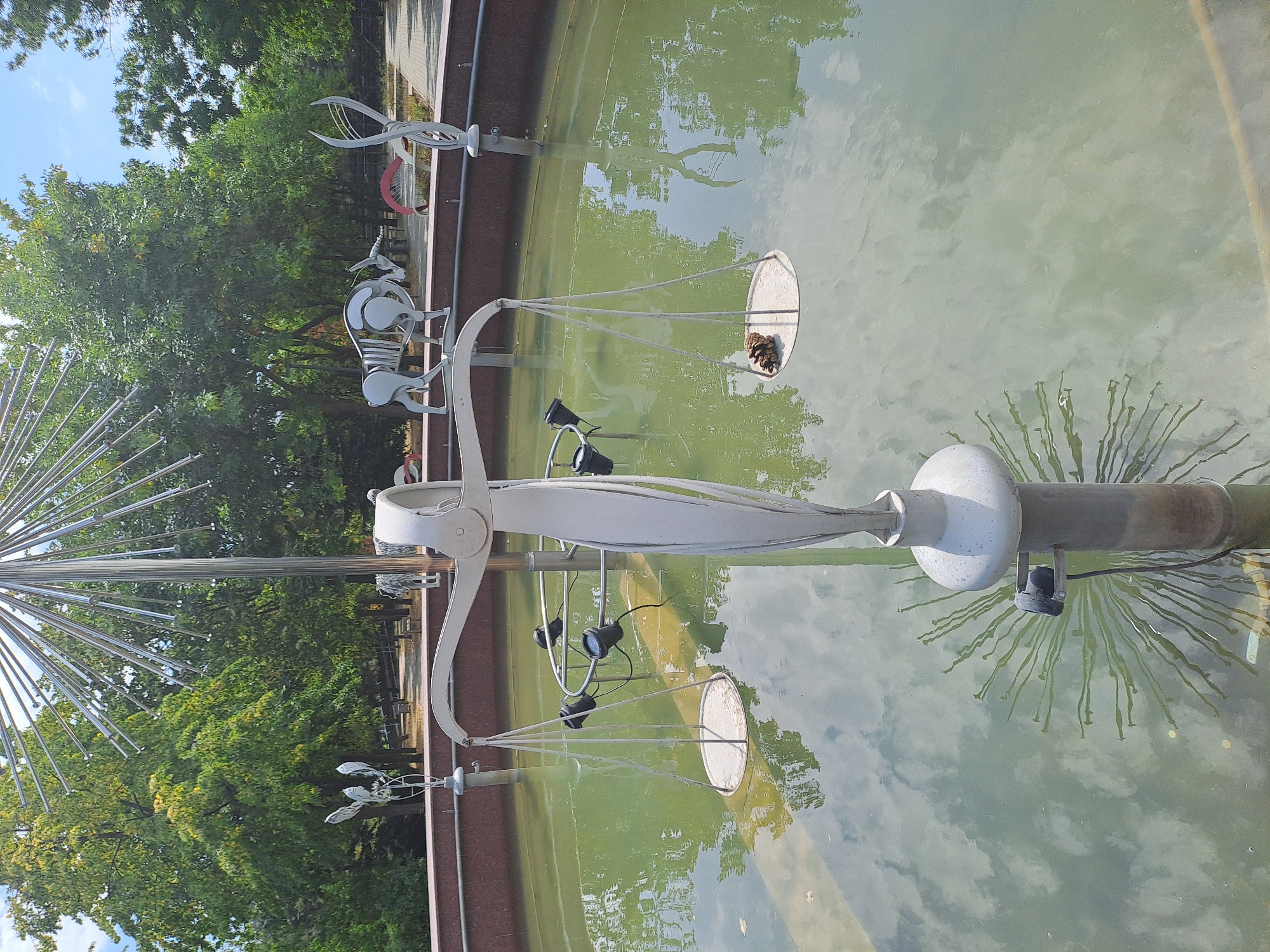 The image shows a fountain sculpture in a park. The sculpture is shaped like a set of scales, with a figure standing in the center. The scales have two pans, one of which contains a pine cone. The fountain is filled with green water, and the reflection of the trees and sky is visible on the surface. In the background, there are several other sculptures of animals, including what appears to be a zebra and an antelope. There are also trees and a paved walkway visible in the background. The overall scene suggests a public park or garden area with artistic installations.
This fountain is located in the Dendrarium Park in Chișinău, Moldova. The park is a large botanical garden that features a diverse collection of trees and plants, as well as several sculptures and art installations. This particular sculpture is a representation of the constellation Libra, and it is a popular attraction in the park.