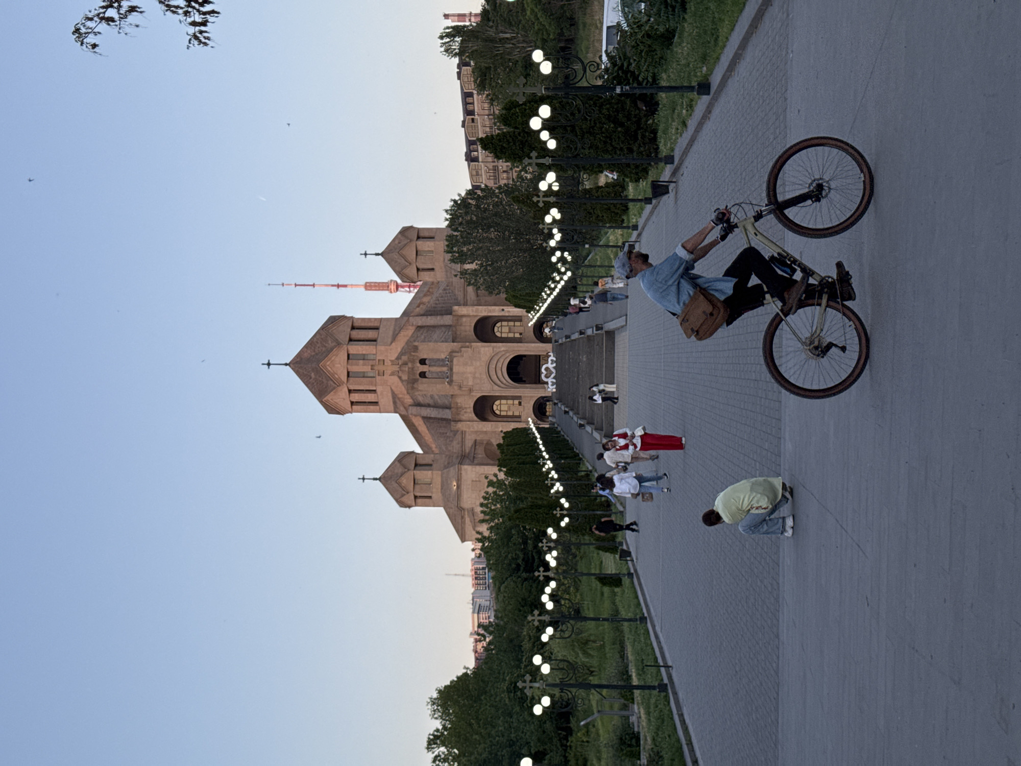 The image depicts the Saint Gregory the Illuminator Cathedral complex in Yerevan, Armenia. The sandstone cathedral, identifiable by its pointed rooflines and crosses, is the focal point, positioned on the left of the image.
To the right of the cathedral, a wide, paved walkway is visible, adorned with regularly spaced lampposts. Several individuals are scattered along the walkway. One person is performing a stunt on a bicycle. Others are walking or standing.
The background includes buildings and foliage, typical of an urban environment. The sky transitions from a pale blue near the horizon to a lighter tone overhead. The image appears to have been rotated.