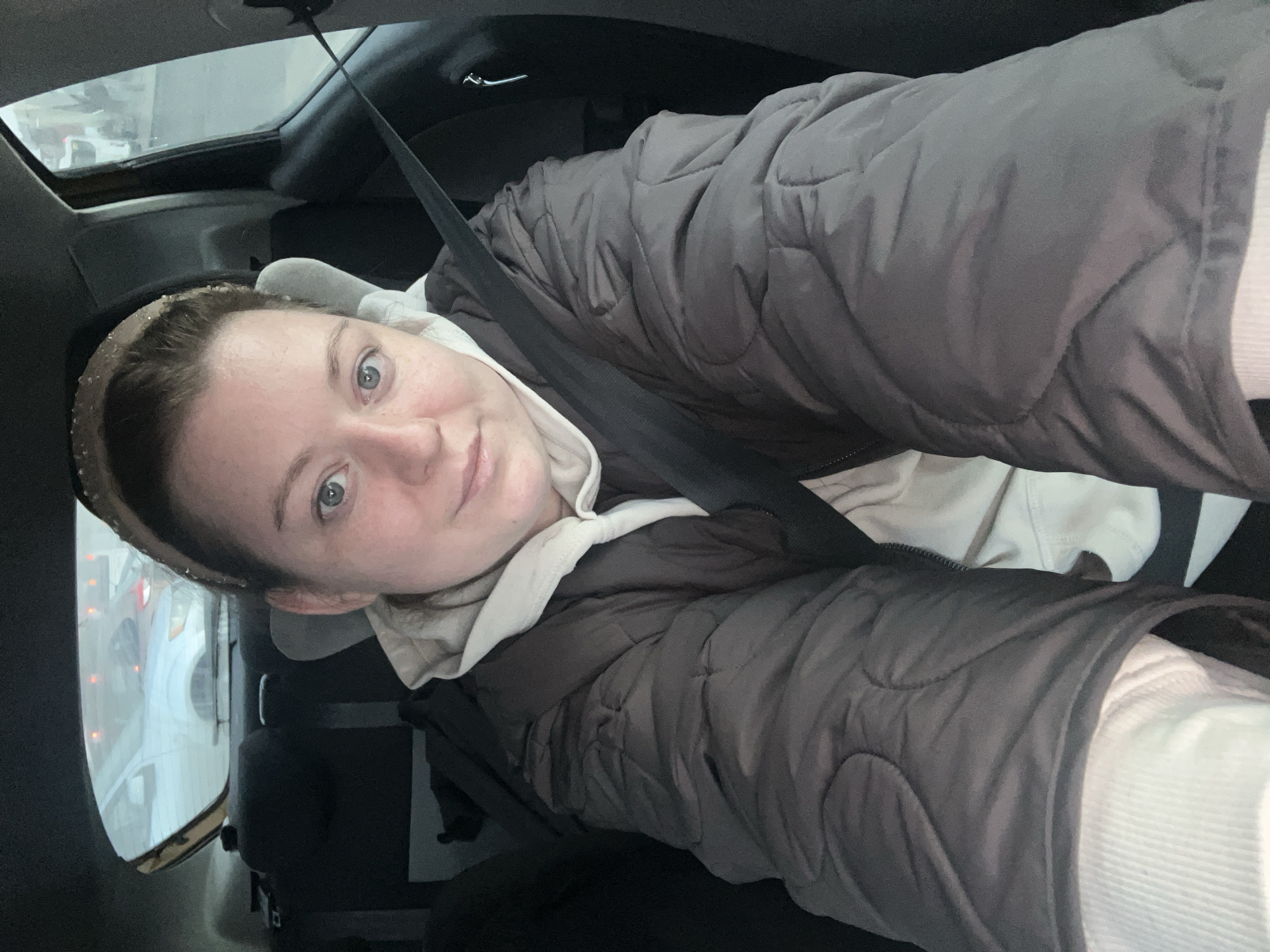The image shows a person in a car, presumably the driver given the angle and presence of a seatbelt. The person has fair skin, blue eyes, and is wearing a neutral-toned headband, a light-colored hoodie, and a dark gray quilted jacket. The background seen through the car windows includes other vehicles and what appears to be an urban environment, potentially aligning with the reported location of Klin, Russia. The sunroof of the car is partially visible.