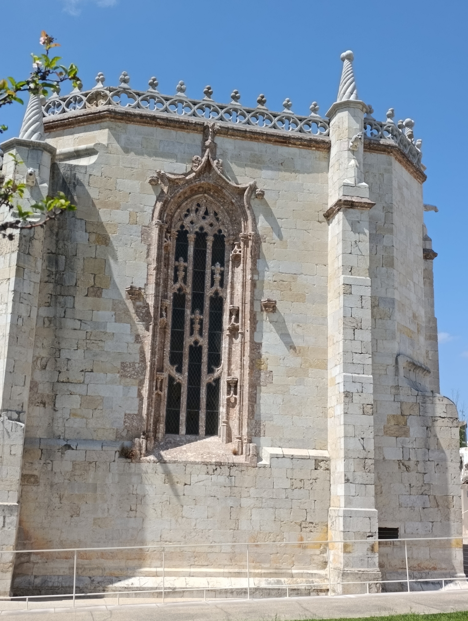 A stone building with a tall, ornate window and a decorative roofline is shown under a clear blue sky in Setúbal, Portugal. The building's exterior is constructed from light-colored stone blocks, with visible texture and variations in hue. The window is Gothic in style, featuring intricate tracery and multiple panes. The roofline is adorned with delicate stonework and spire-like finials. A few small, green branches with pink flowers are visible in the upper left corner of the image, suggesting the presence of a tree or shrub. The lower part of the building is bordered by a white railing and a paved area. The lighting suggests it is daytime, with distinct shadows cast on the building's facade.