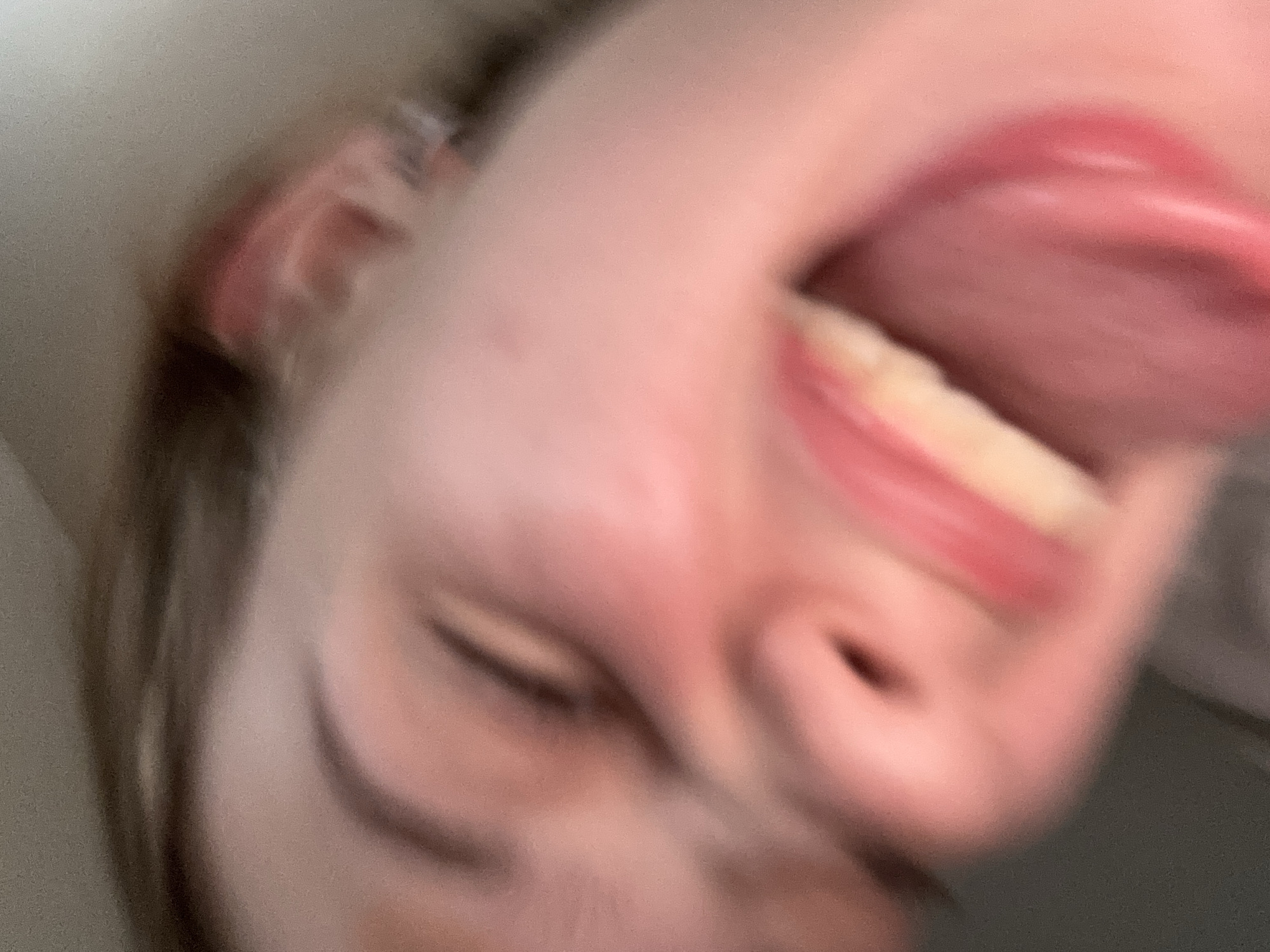 A close-up, blurred photograph captures a person's face from a low angle. The individual's eyes are closed, and their mouth is open, with their tongue extended. The skin is flushed, and there appears to be some makeup on the lips. The background is out of focus and appears to be a light-colored surface. No specific location in the city of Unknown, Russia can be determined from the image.