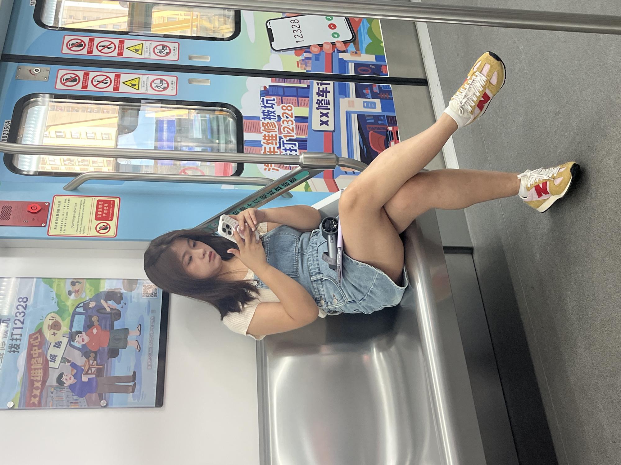 A young East Asian woman with dark hair is sitting on a metal bench on a subway car, looking at her smartphone. She is wearing a light-colored short-sleeved shirt and a denim overall skirt, with white socks and colorful sneakers. Her legs are crossed. To her left, a large poster on the wall displays illustrations of people and a car, along with Chinese text and a phone number. To her right, the subway car doors are decorated with bright blue panels featuring a cityscape mural and various safety warnings and instructions in Chinese. The environment is clean and well-lit, typical of public transportation in an urban setting. The scene appears to be in the city of Tiaodeng, China, based on the visual cues and implied location.