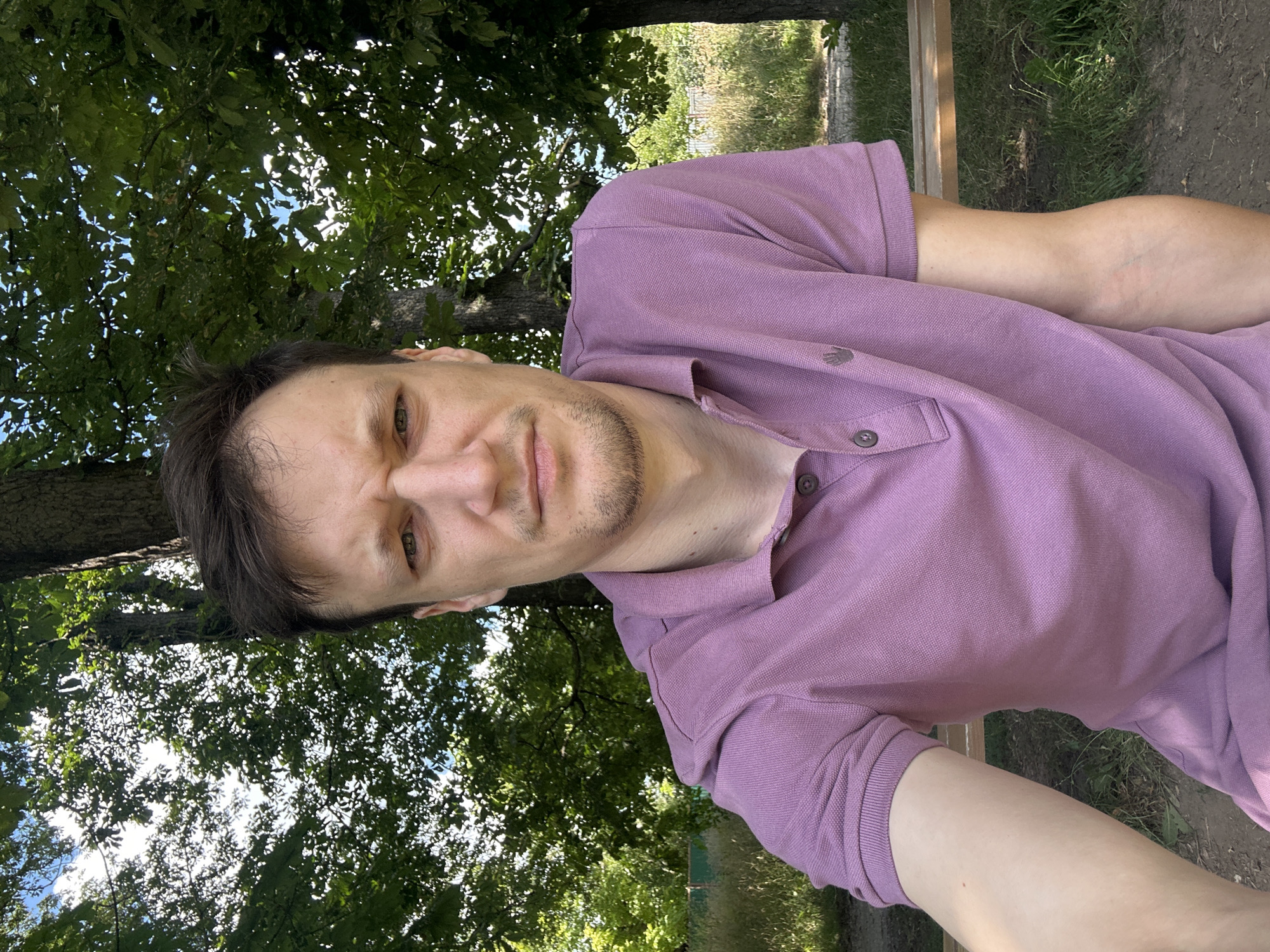 The image is a selfie taken outdoors, likely in Makiv, Ukraine, with a focus on a man sitting on a park bench. The man, with short brown hair and a slight stubble, is wearing a purple polo shirt. He is looking directly at the camera with a furrowed brow. Behind him, lush green trees with dappled sunlight filtering through the leaves create a natural, shaded environment. The ground appears to be dirt and grass. The overall scene suggests a casual outdoor setting on a sunny day.