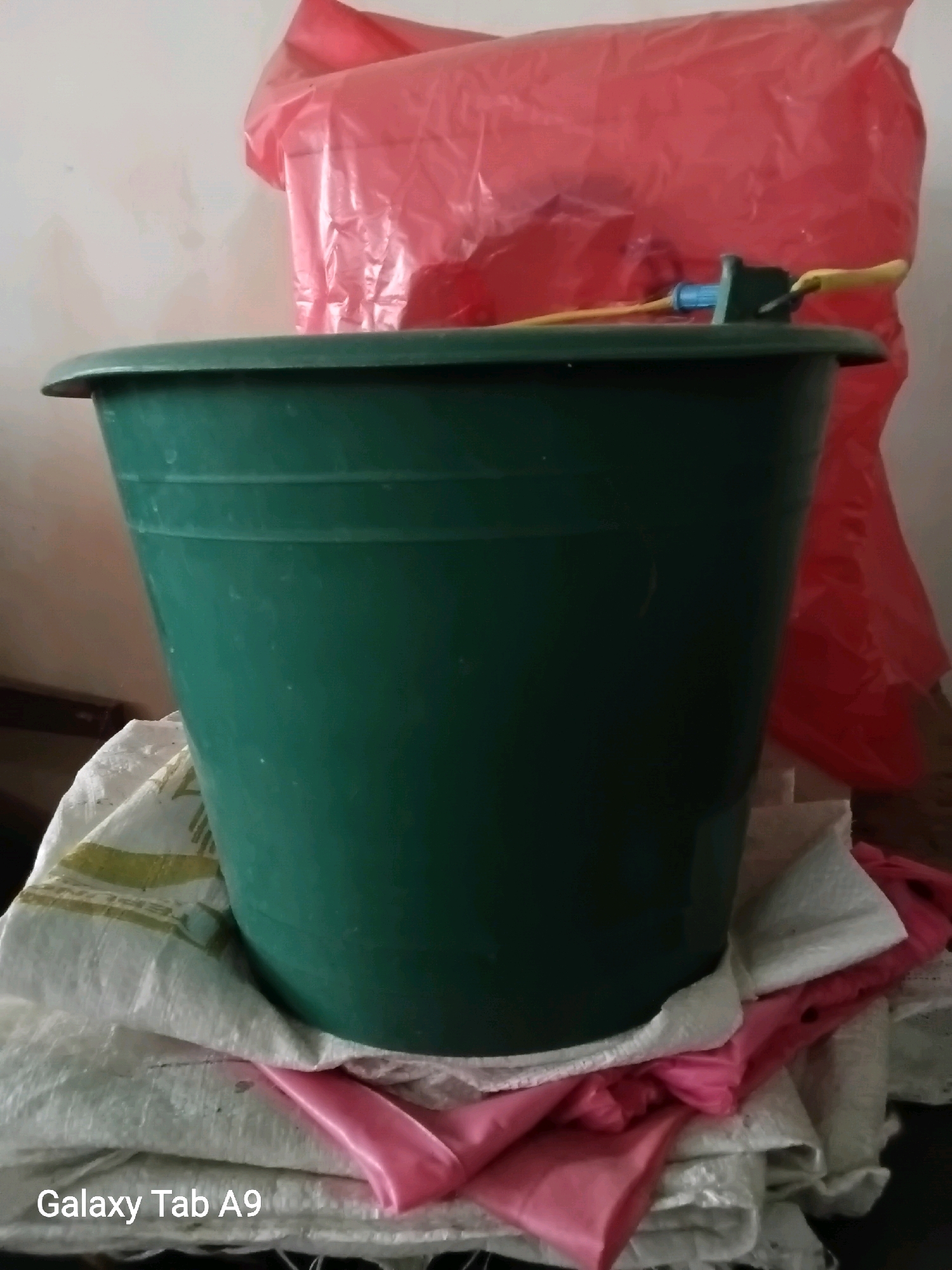A dark green plastic bucket is positioned in the foreground of the image, resting on a pile of white and pink fabric. Behind the bucket, a large, crinkled red plastic bag is visible, partially obscuring the background. A small blue and green faucet-like object is attached to the rim of the bucket, with a yellow rod extending from it. The overall scene appears to be indoors, with a plain, light-colored wall in the background. The lighting is somewhat dim, creating soft shadows. The bottom left corner of the image displays the text "Galaxy Tab A9".