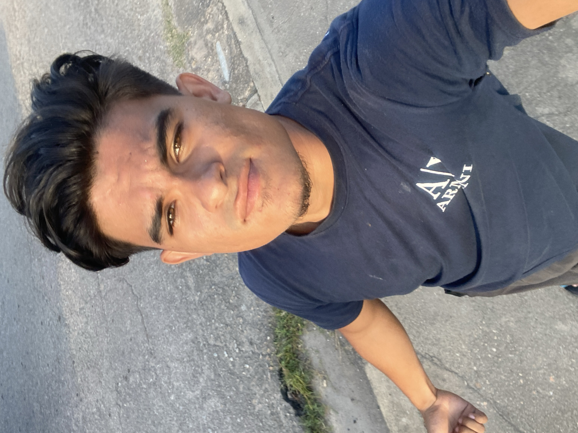 A young man stands on a concrete surface, likely a sidewalk, in an urban setting. He is wearing a navy blue t-shirt with a white "Armani" logo. The man has dark hair styled with a wave in the front and a sparse goatee. The location is possibly Juan Griego, Venezuela, as per the request, but there is no visible signage or landmarks to confirm this. The background consists of gray pavement with some grass growing along the edge. The time of day appears to be either early morning or late afternoon, judging by the angle and quality of light. The man is looking directly at the camera.